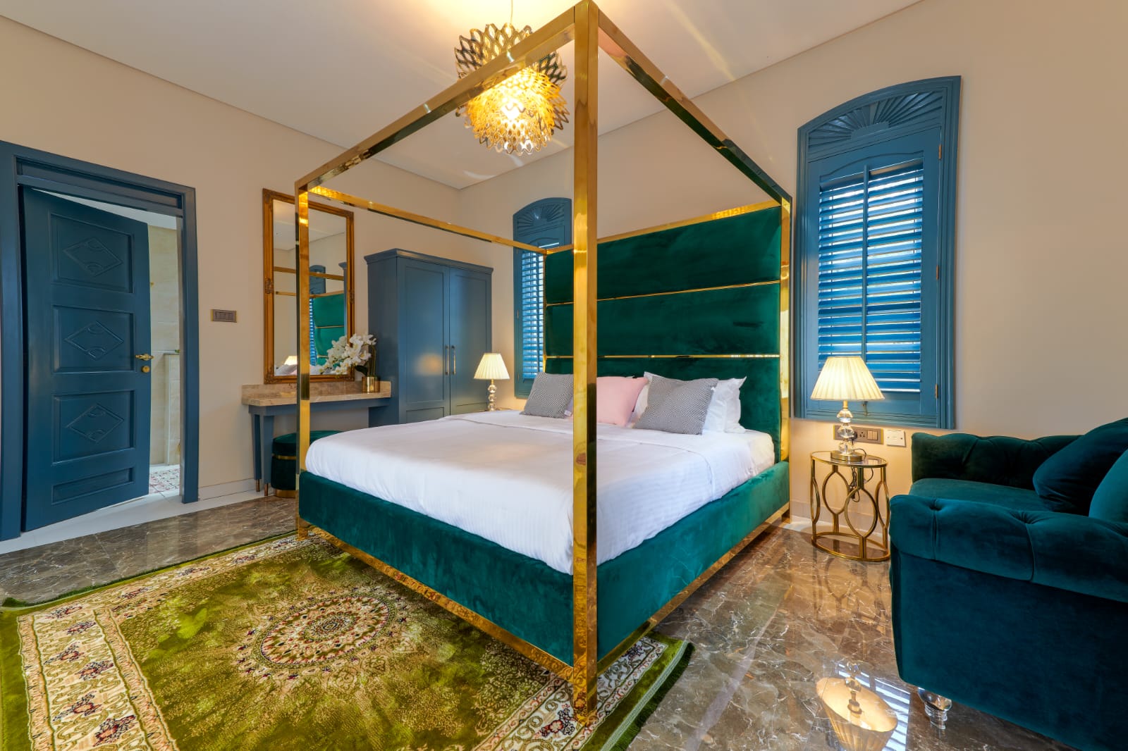 Luxurious bedroom with a green velvet canopy bed featuring gold frame, blue shuttered windows, blue wardrobe, green patterned rug, and matching green armchair.