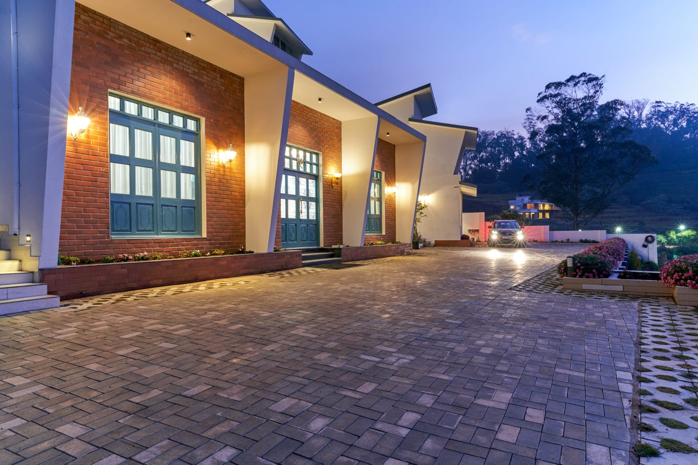 Modern house exterior at dusk with brick walls, lit porch lights, paved driveway, and a car with headlights on.