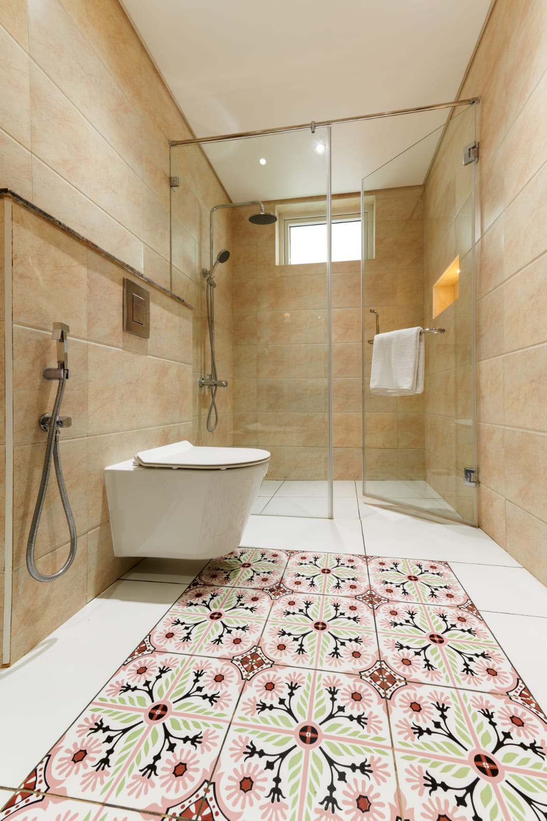 Modern bathroom with beige tiled walls, wall-mounted toilet, patterned floor tiles, and glass-enclosed shower area with a towel on the rack.
