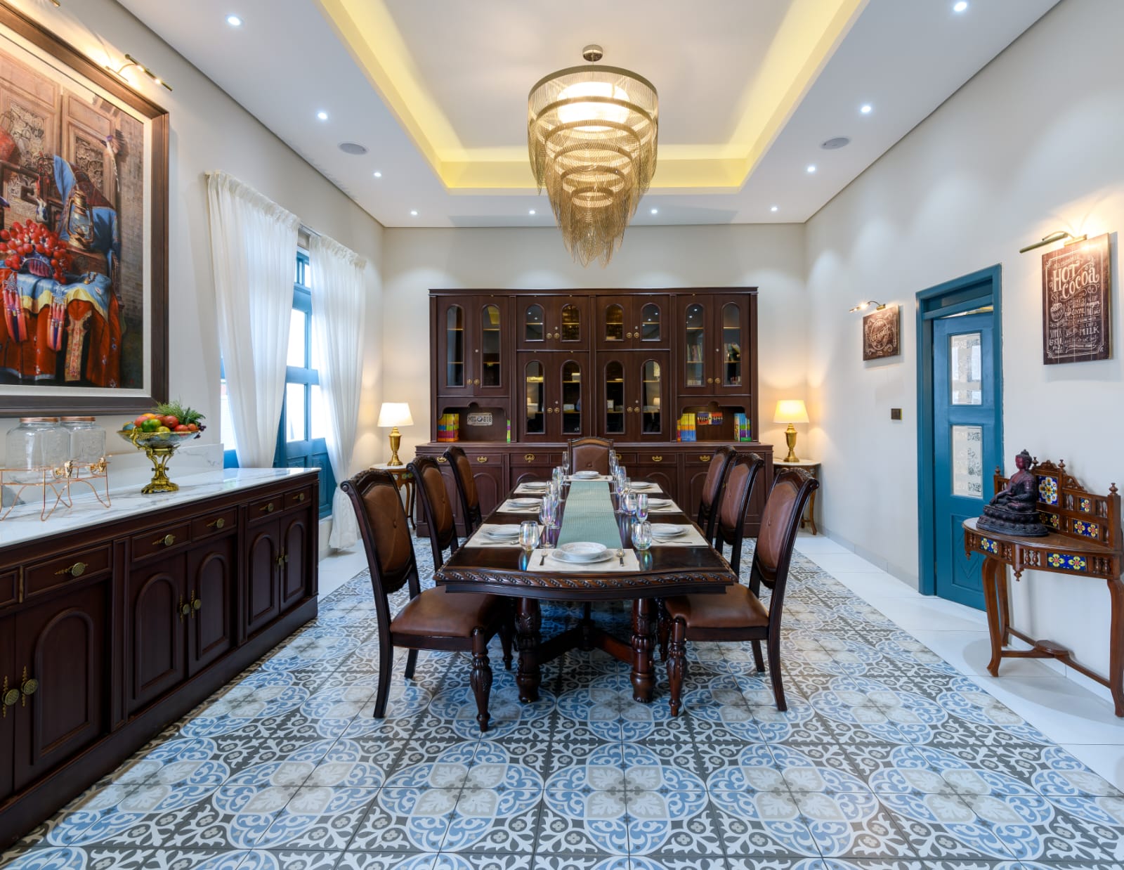 Elegant dining room with a long dark wooden table set for eight, ornate blue and gray patterned floor tiles, classic wooden cabinets, and a large modern chandelier.