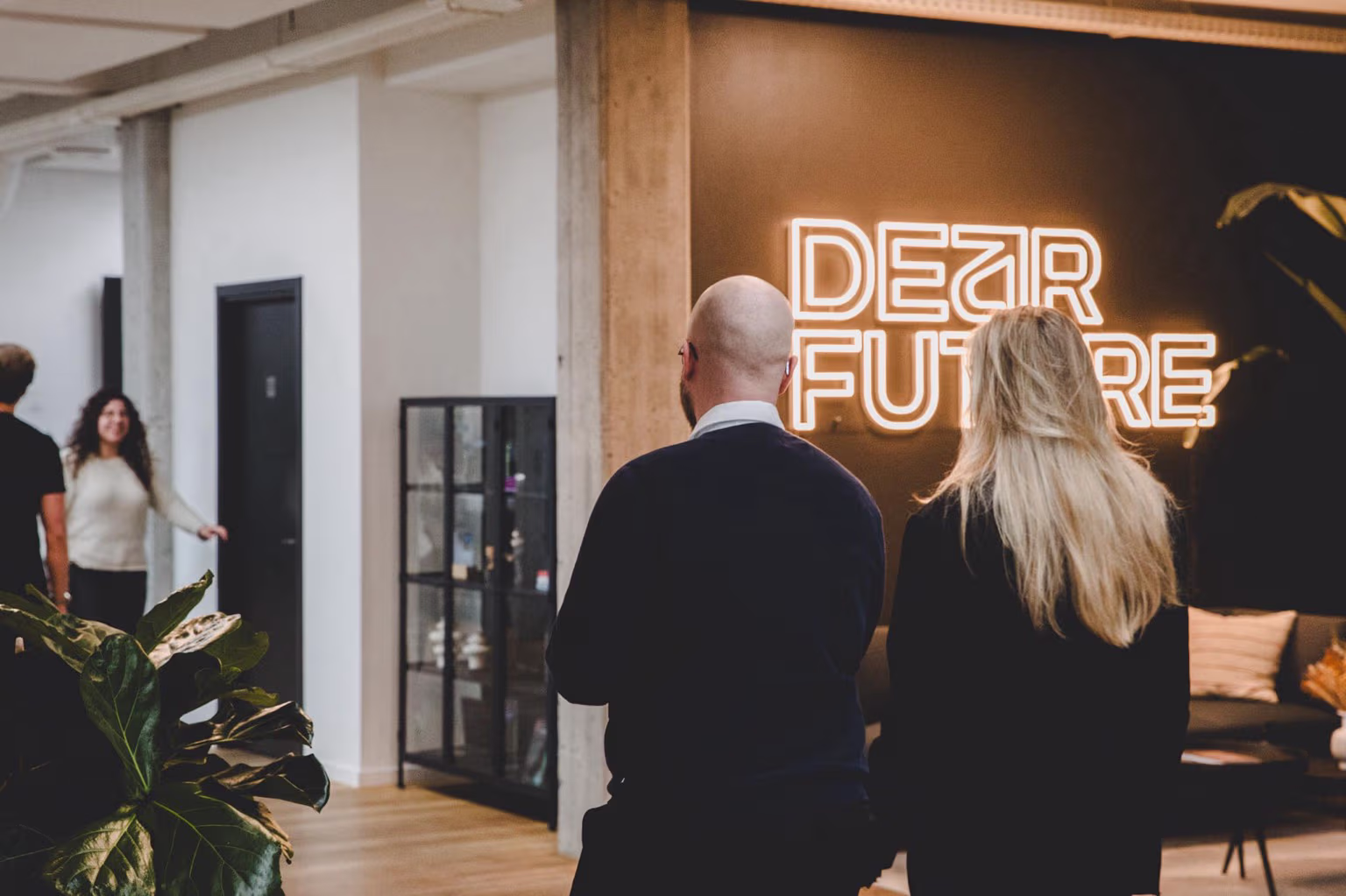 To personer set bagfra i en moderne kontorindgang med en neonlys-skilt, der siger 'DEAR FUTURE'.