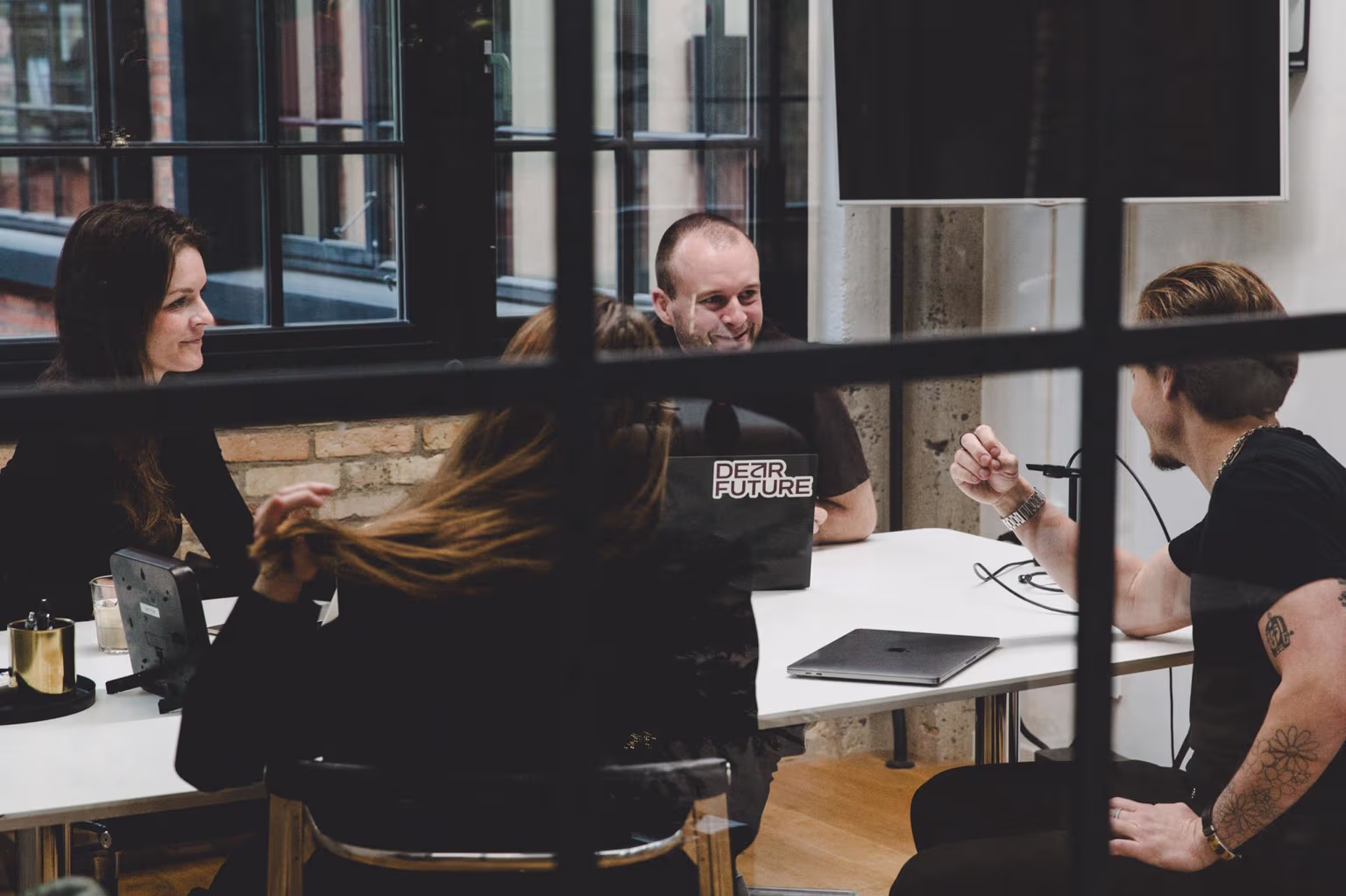 Fire personer i en kontorsituation, der sidder rundt om et bord og diskuterer, med en bærbar computer med teksten 'Dear Future'.