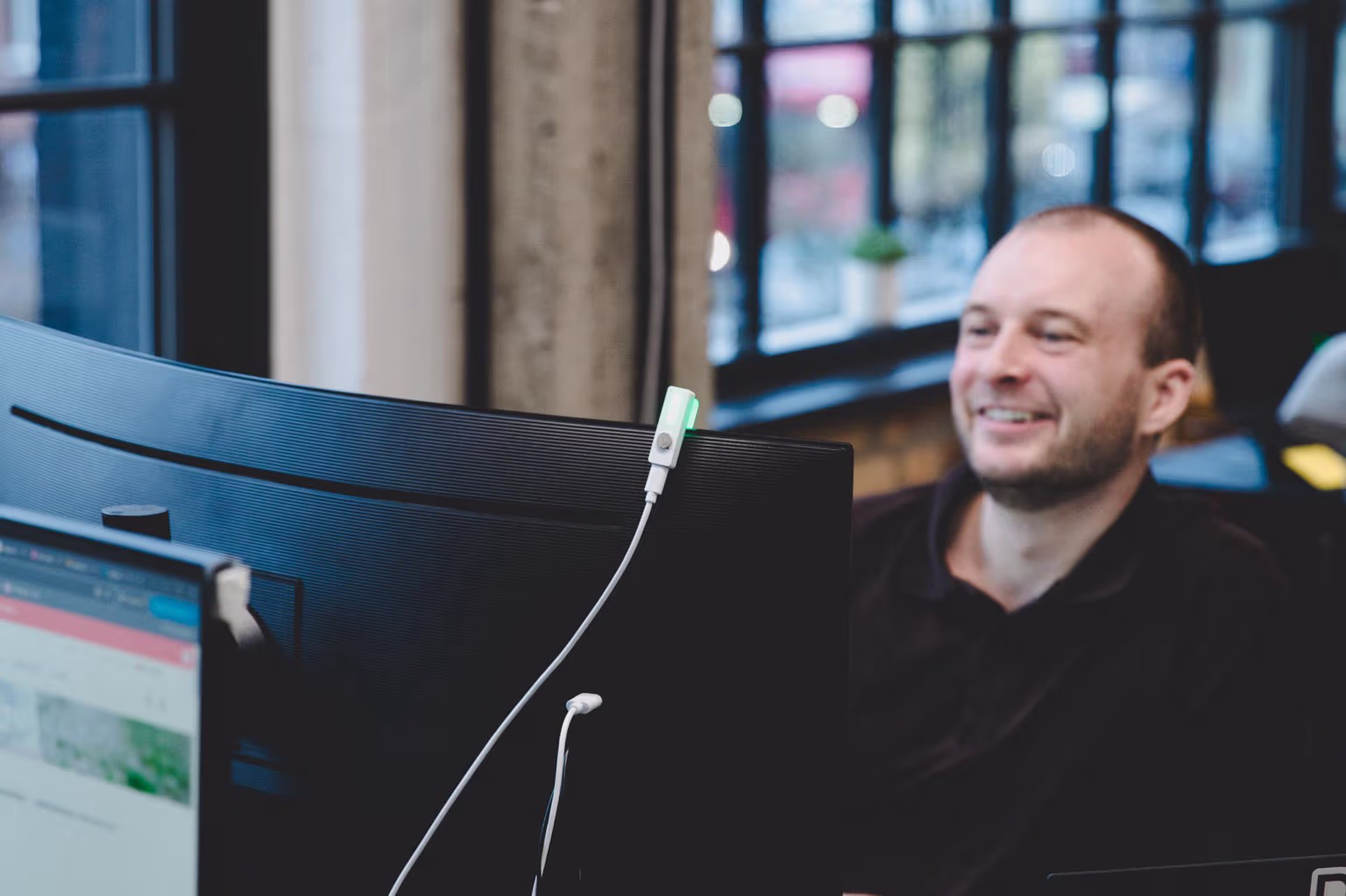 Mand smilende ved sit skrivebord med en computerskærm med tilsluttet webcam i et moderne kontormiljø.