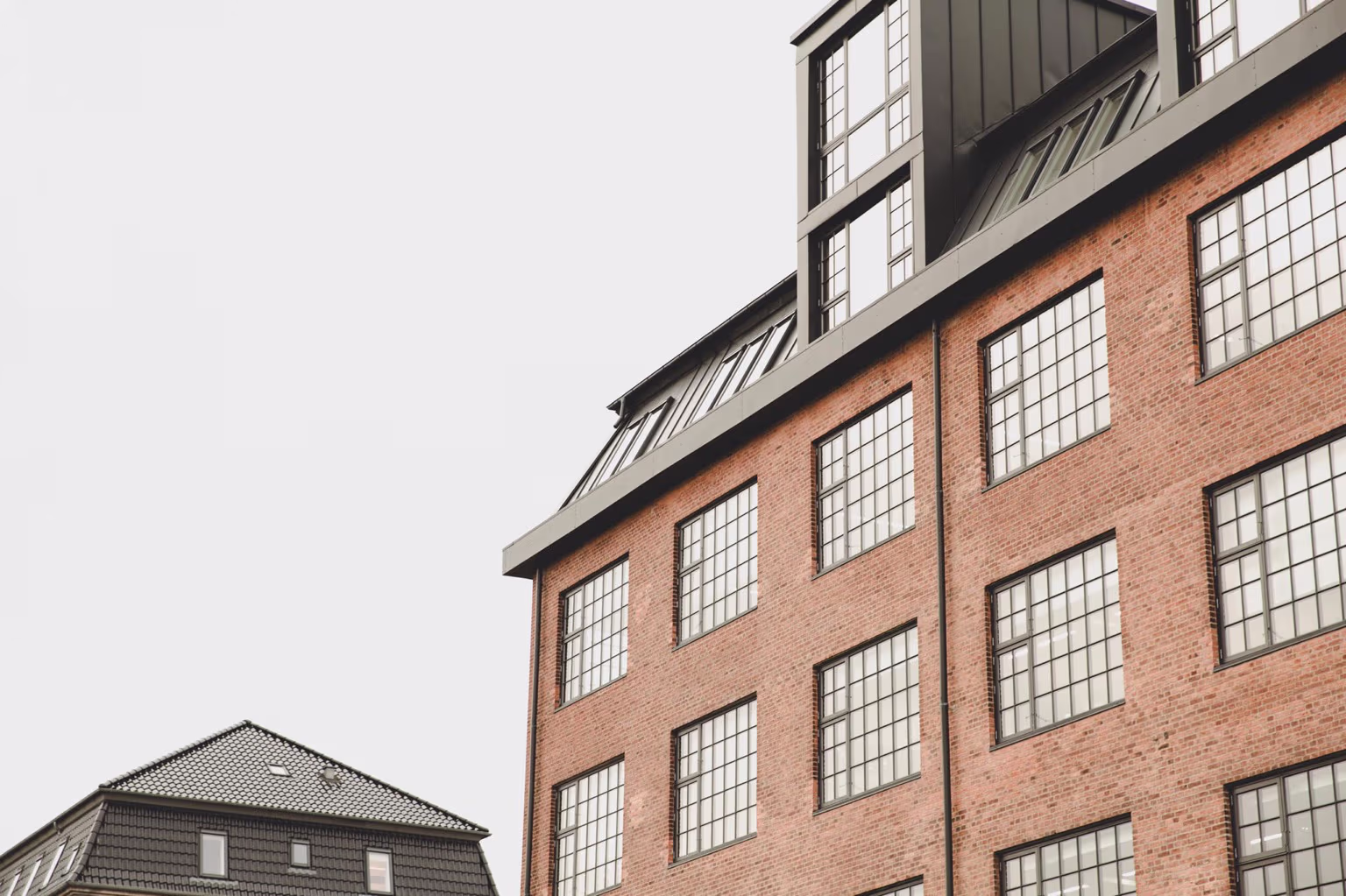 Rød murstensbygning med mange ruder og en tagkonstruktion med sorte kviste mod en grå himmel.