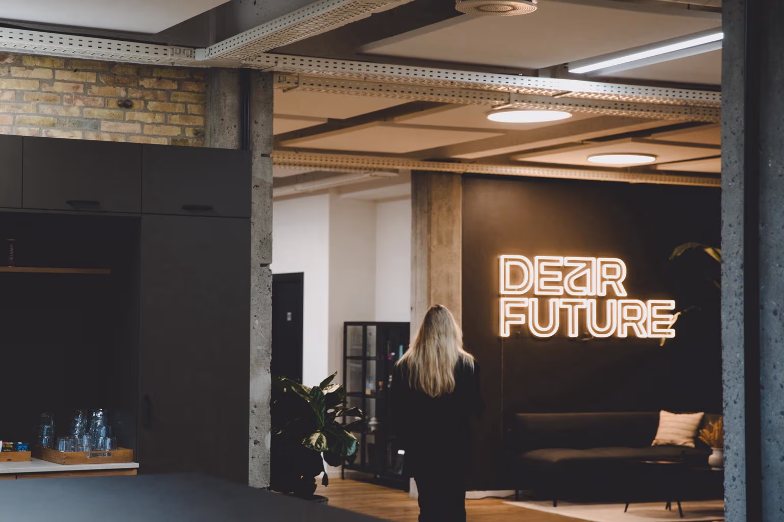 En kvinde med langt lyst hår går mod en væg med neonskiltet 'DEAR FUTURE' i et moderne kontormiljø.