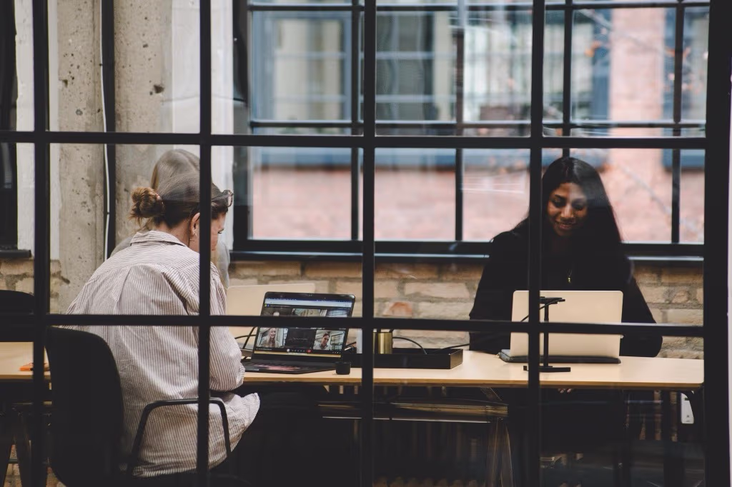 To personer arbejder på bærbare computere ved et bord i et lyst rum med mursten og store vinduer.