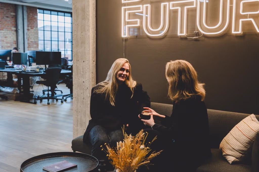 To kvinder sidder ved en sofa i et moderne kontor, den ene holder en kop, og et neonskilt med ordet FUTURE lyser i baggrunden.