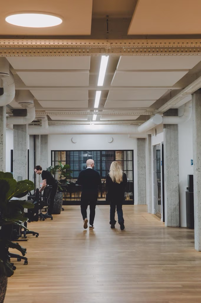 To personer går ned ad en lys gang i et moderne kontor med store vinduer og trægulv.