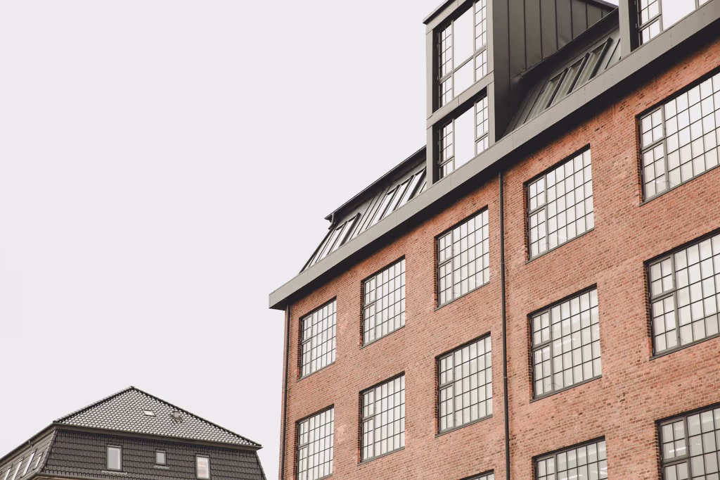 Brun murstensbygning med store firkantede vinduer og tagvinduer under en grå himmel.