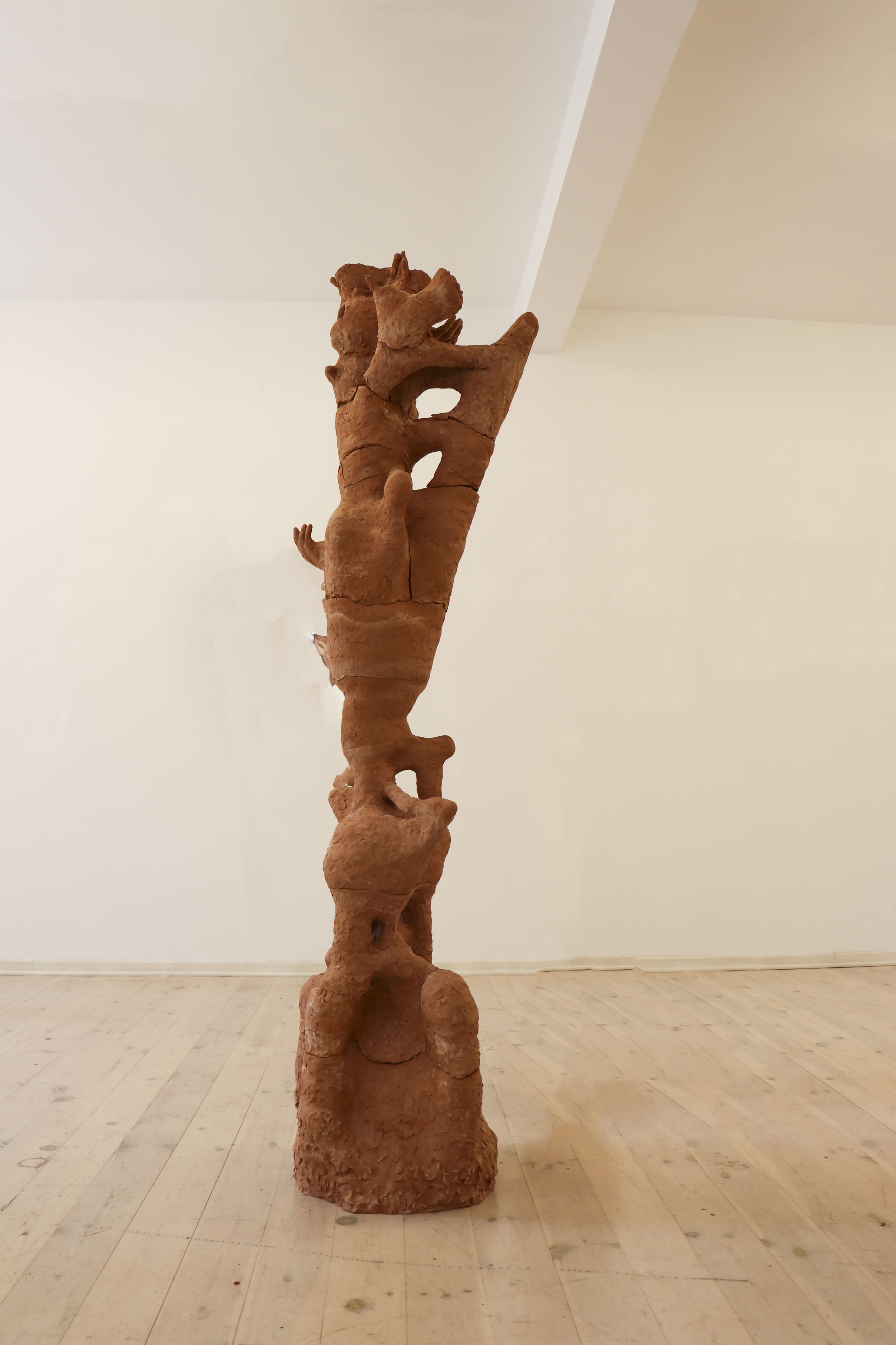 Abstract tall reddish-brown clay sculpture with irregular shapes and openings, displayed in a gallery with wooden floor and plain walls.