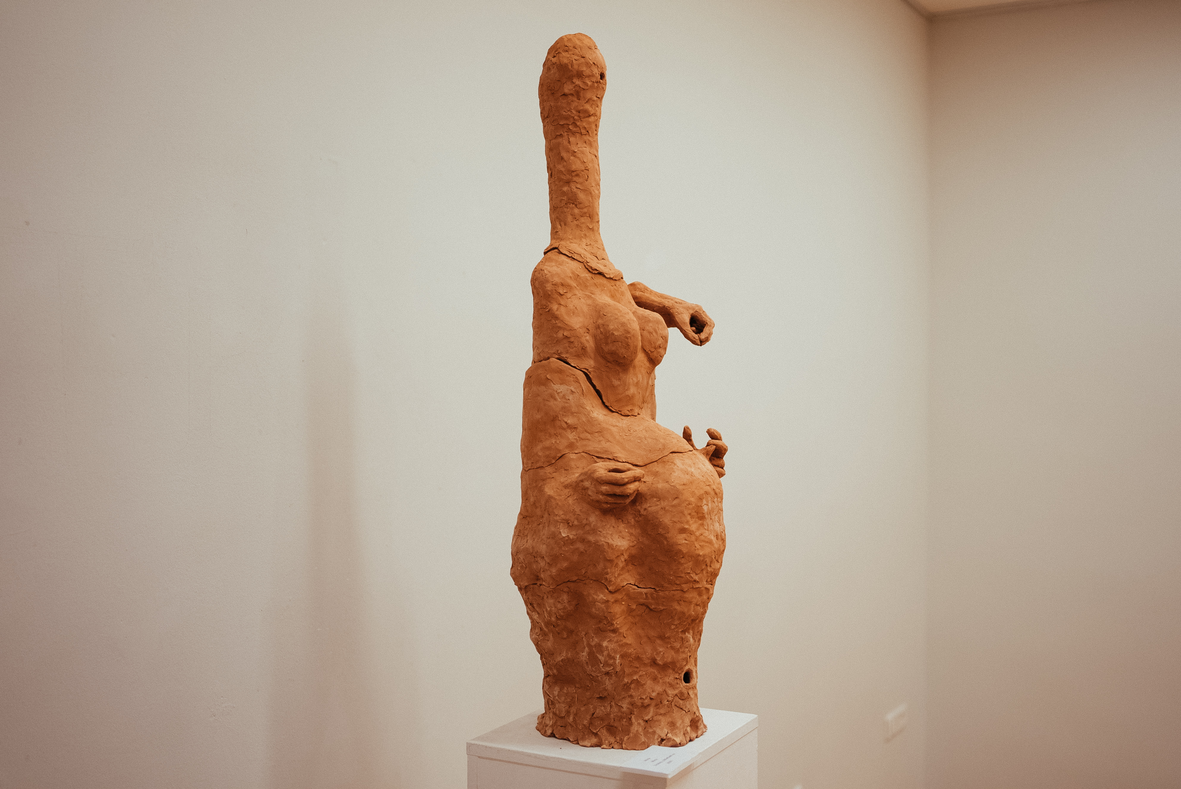 Abstract terracotta sculpture of a female figure with exaggerated pregnant belly and elongated neck.