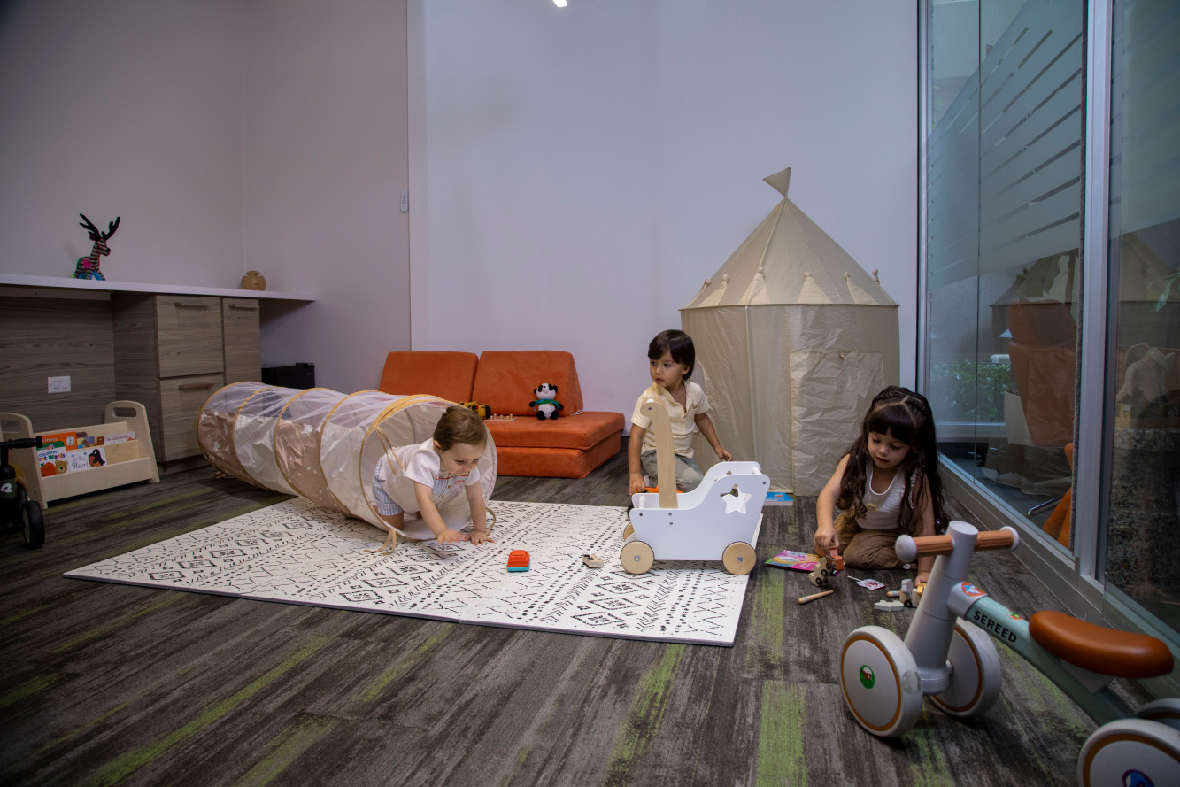 Three children playing in a bright playroom with a toy tunnel, wooden baby walker, beige play tent, and various toys scattered on the rug and floor.