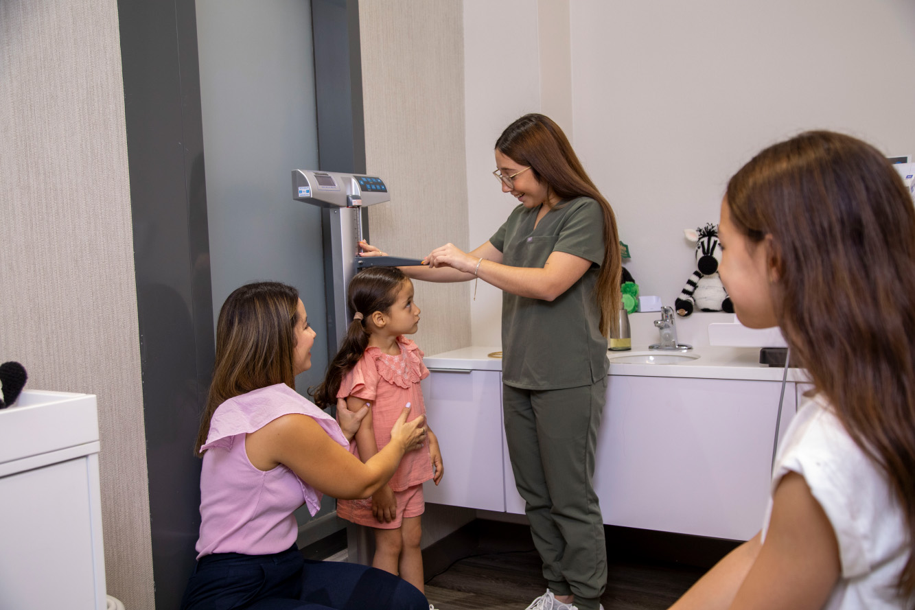 A healthcare professional measures the height of a young girl while her mother supports her, another child watches nearby.