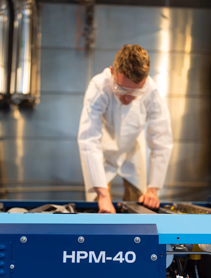 Person in white lab coat and safety glasses working with machinery labeled HPM-40 in an industrial setting.
