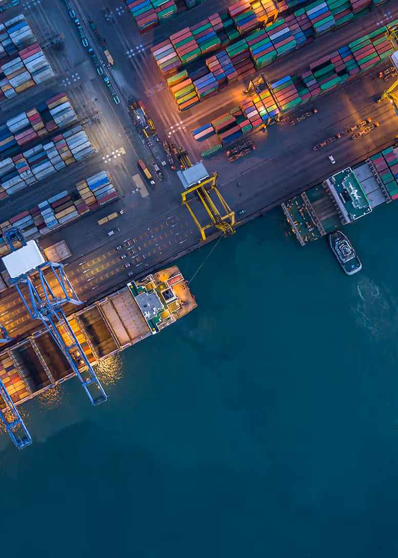 Vue aérienne d'un port maritime très actif avec des conteneurs colorés empilés, de grandes grues, des cargos à quai et une eau bleue calme.