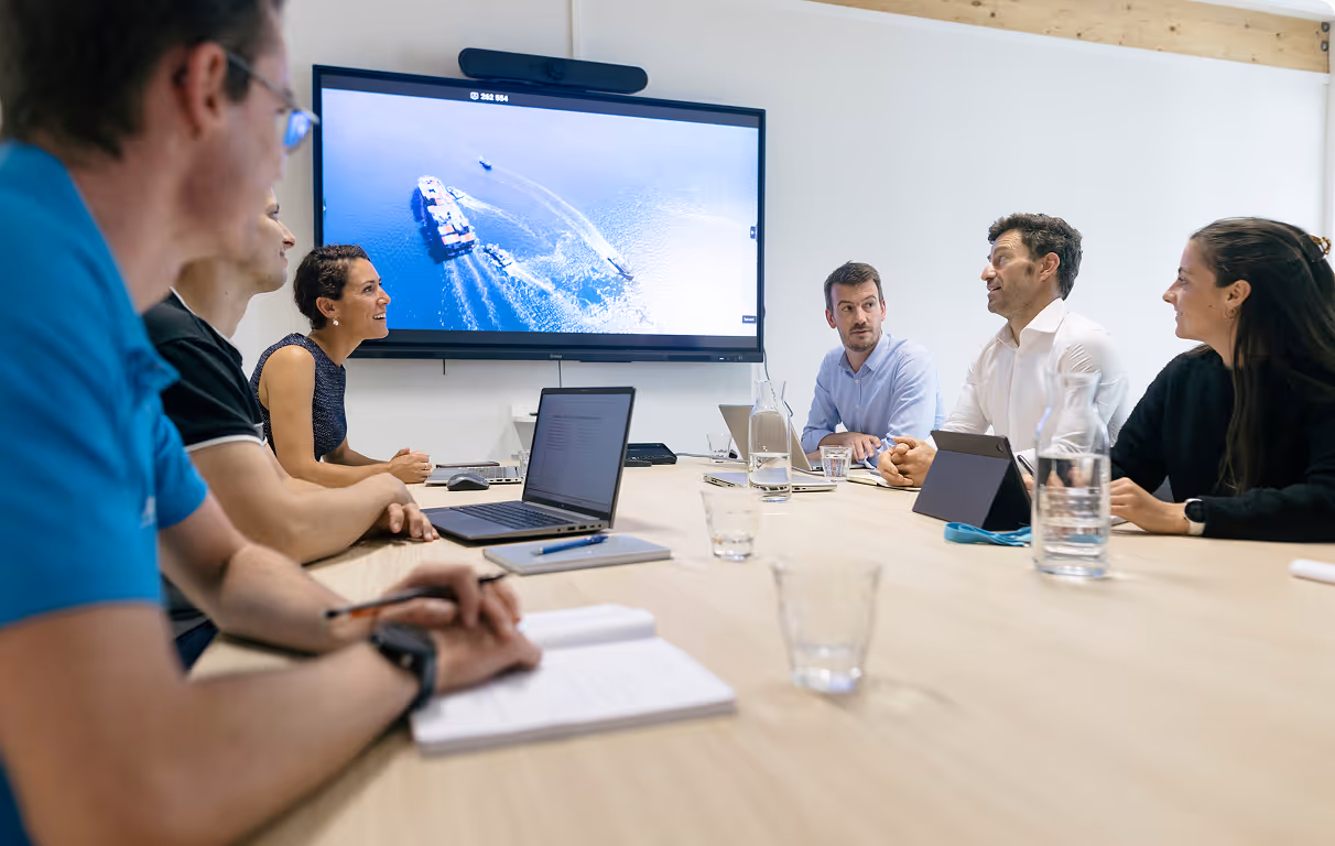 Six collègues discutent dans une salle de réunion, tandis qu'un grand écran affiche une vue aérienne de bateaux sur l'eau.