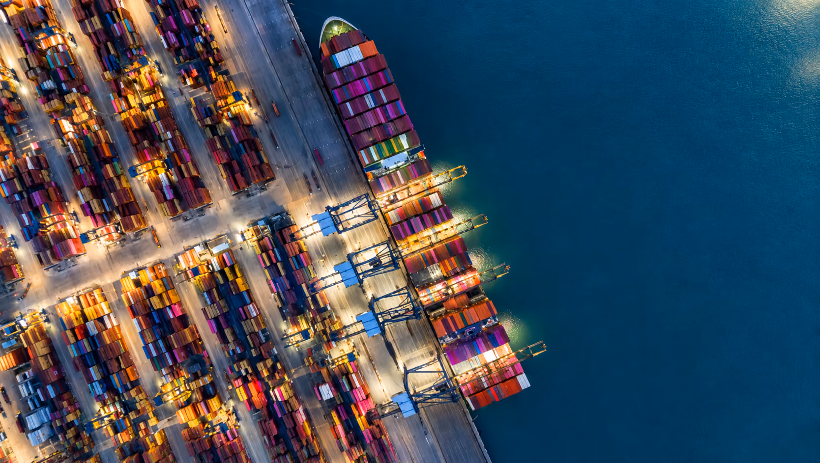 Aerial view of a busy shipping port with colorful stacked containers, large cranes, docked cargo ships, and calm blue water.