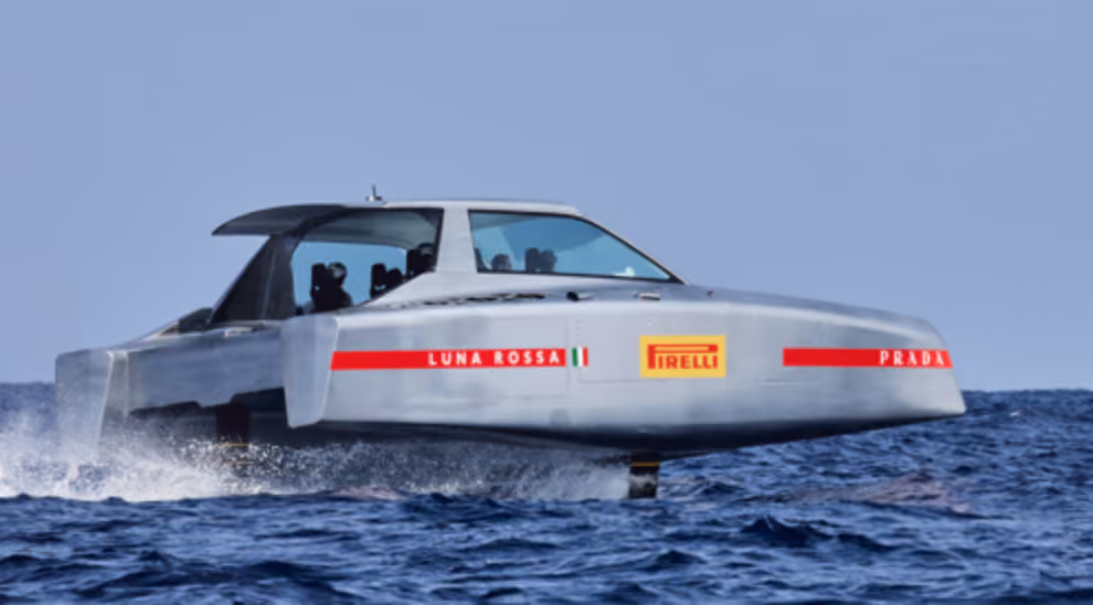 Silver Luna Rossa foil boat with Pirelli and Prada logos hydrofoiling above ocean water.