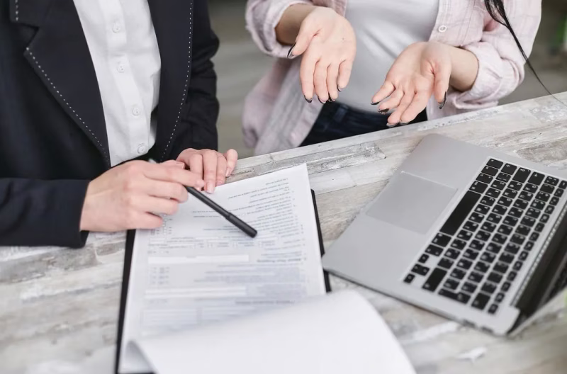 Two people at a table, one pointing a pen at a document, the other gesturing with open hands near a laptop.