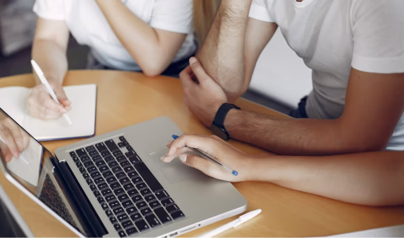 Three people around a laptop at a table, one writing notes while another points on the laptop touchpad.