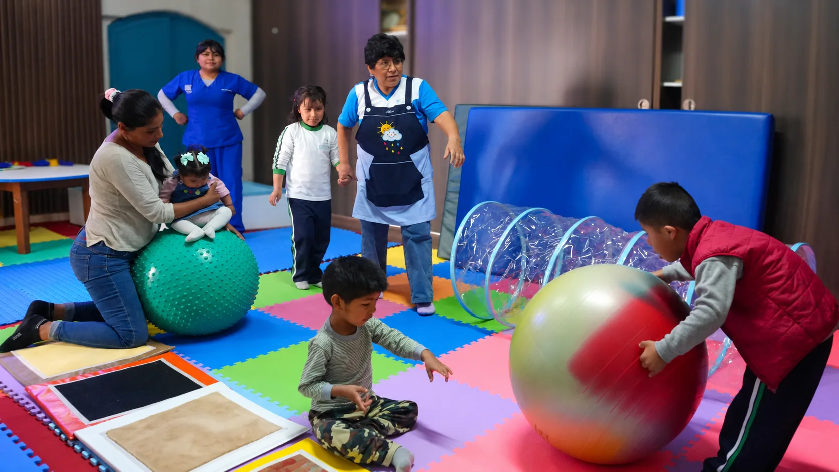 Kinder spielen mit Therapiebällen und einem durchsichtigen Krabbeltunnel auf bunten Bodenmatten, begleitet von einer Mutter und zwei Betreuerinnen