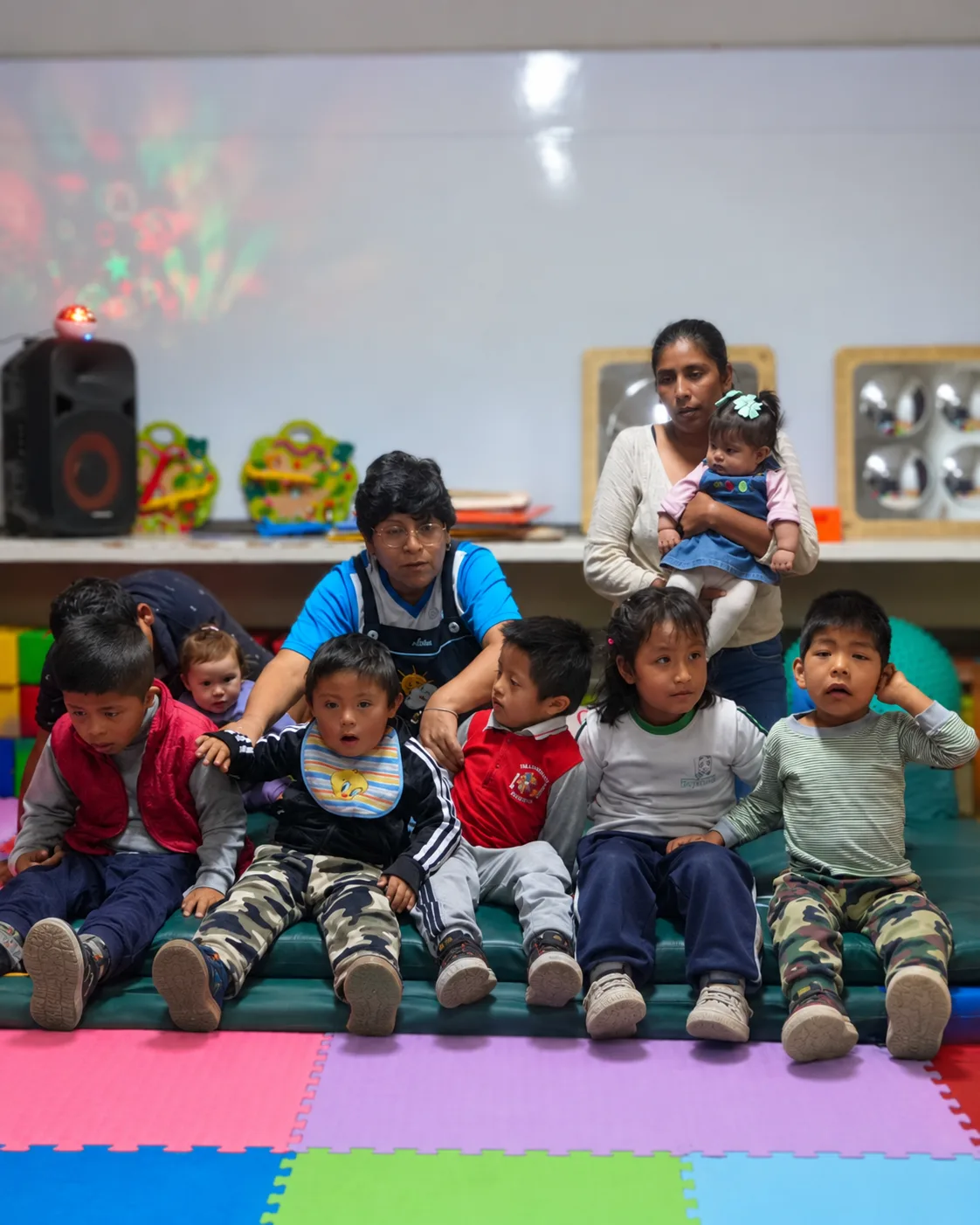 Kinder sitzen nebeneinander auf einer grünen Turnmatte im Aula Multisensorial, hinter ihnen eine Betreuerin und eine Mutter mit Baby, an der Wand Lichtprojektionen und Spiegel