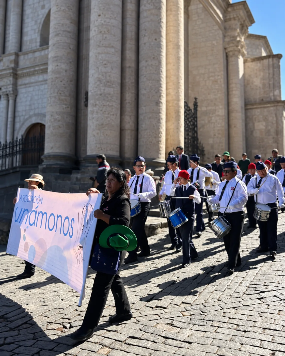 Die Banda von Unámonos in weißen Hemden und dunklen Mützen mit Trommeln vor einer Steinkirche, vorneweg zwei Frauen mit einem Transparent der Asociación Unámonos