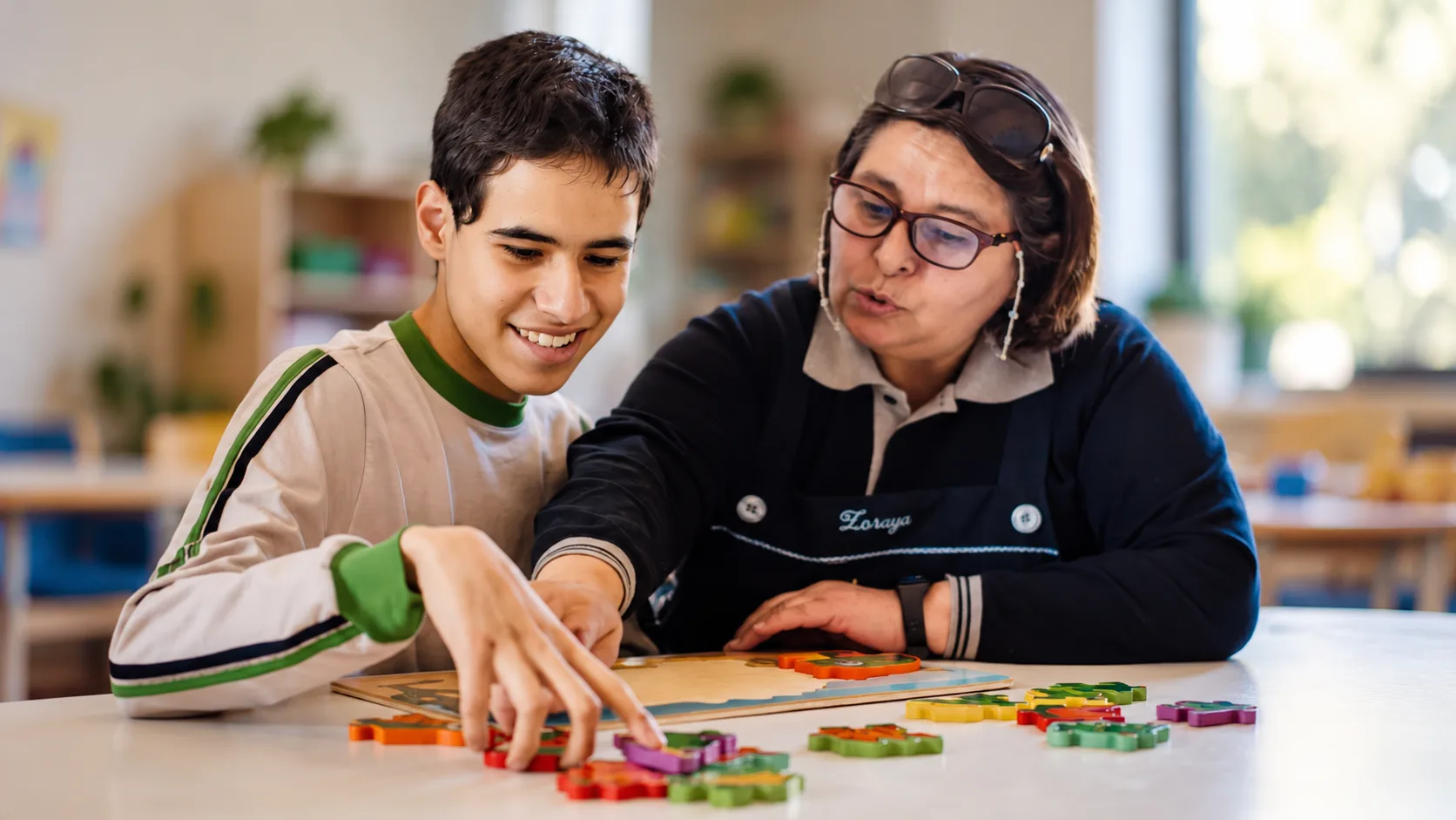 Ein Schüler und eine Mitarbeiterin der Asociación legen gemeinsam ein Holzpuzzle mit bunten Teilen in einem hellen Klassenraum