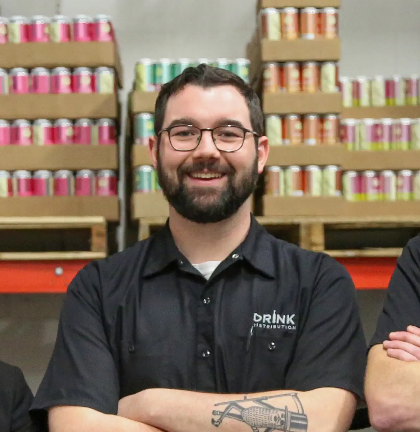 Smiling man with glasses and beard wearing a black Drink Distribution shirt standing with arms crossed in front of shelves filled with colorful canned drinks.