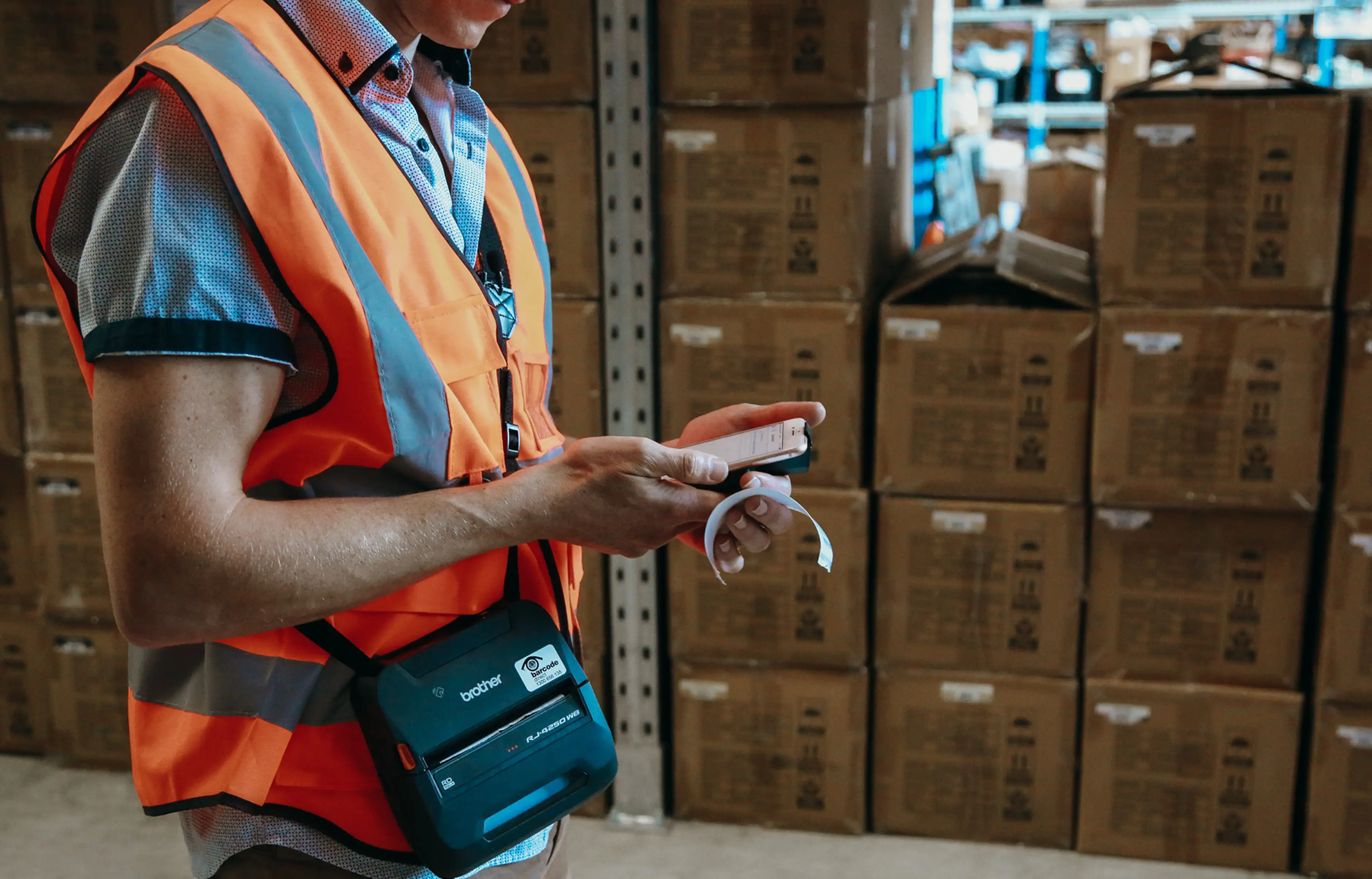 Warehouse worker wearing an orange safety vest using a smartphone and a portable Brother label printer with stacked cardboard boxes in the background.