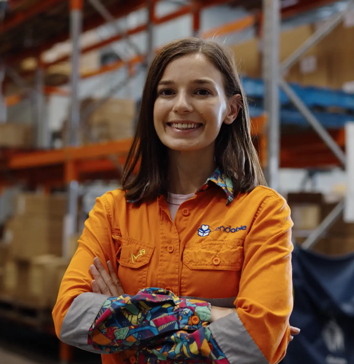 Smiling woman with crossed arms wearing an orange shirt with colorful patterned cuffs in a warehouse.