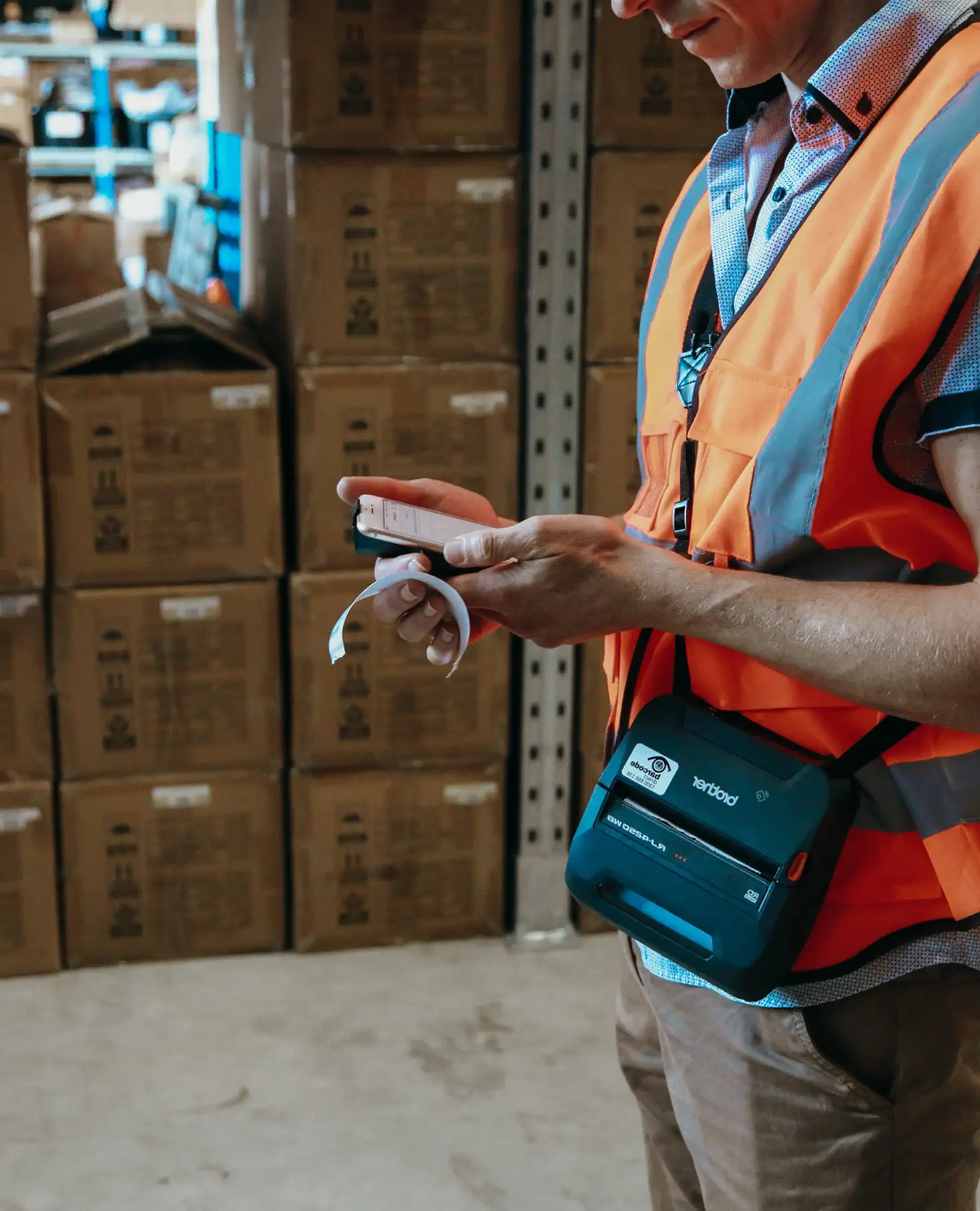 Warehouse worker in orange safety vest holding a mobile device and a printed receipt with stacked boxes in the background.