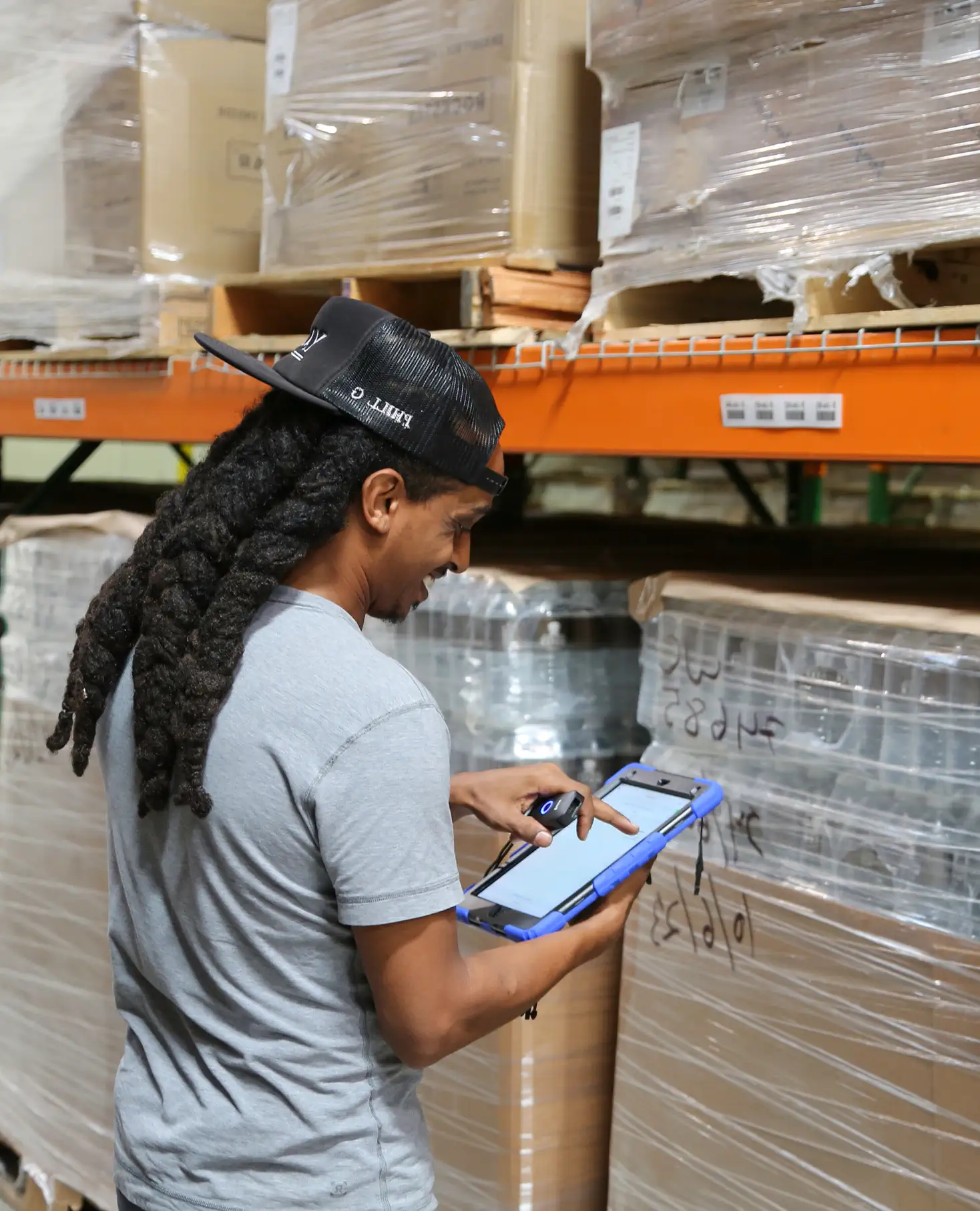 Warehouse worker using a tablet device to check inventory near stacked, shrink-wrapped boxes.