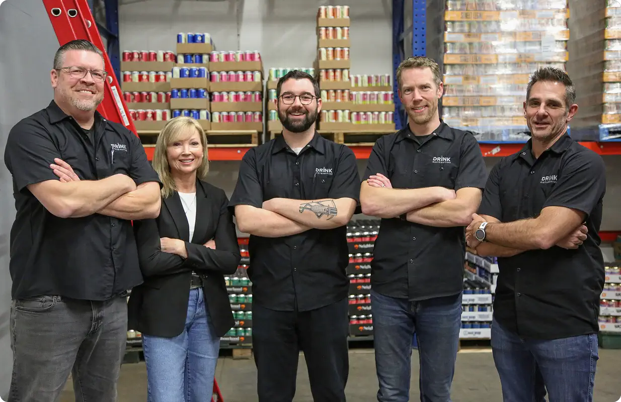 Five people standing with arms crossed in a warehouse with shelves stacked with canned beverages behind them.