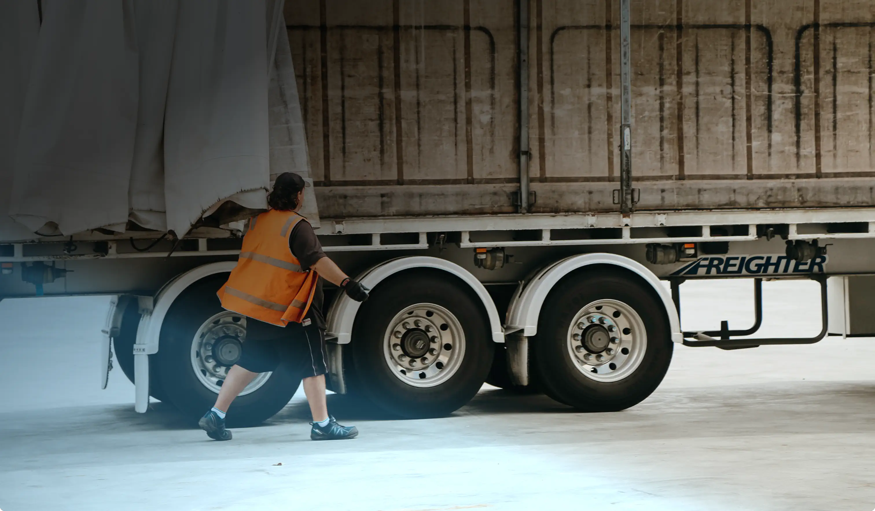 Worker in an orange safety vest walking beside the rear wheels of a large freight truck in a warehouse.