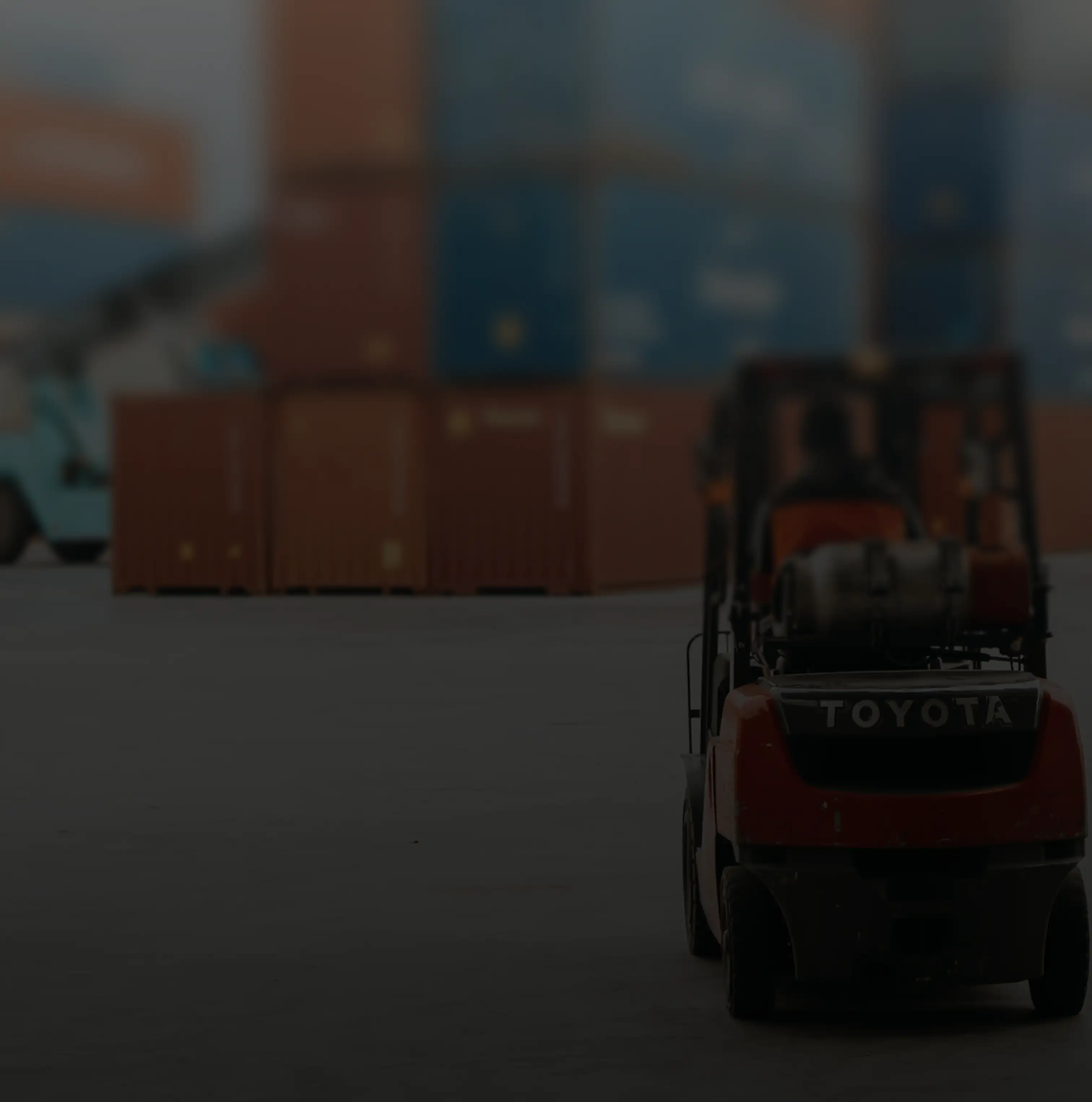 Rear view of a red Toyota forklift moving in an industrial area with stacked shipping containers in the background.