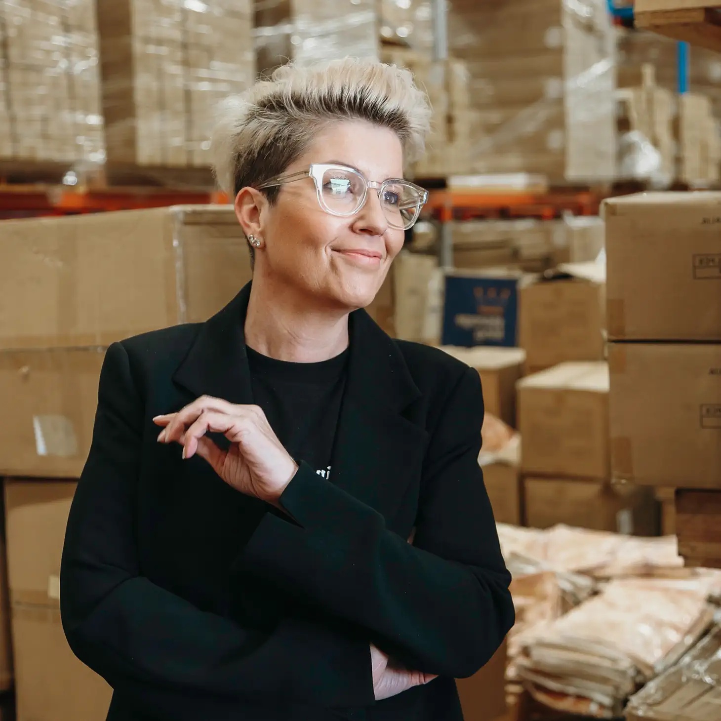 Smiling person with short blond hair and glasses, standing in a warehouse surrounded by stacked cardboard boxes.