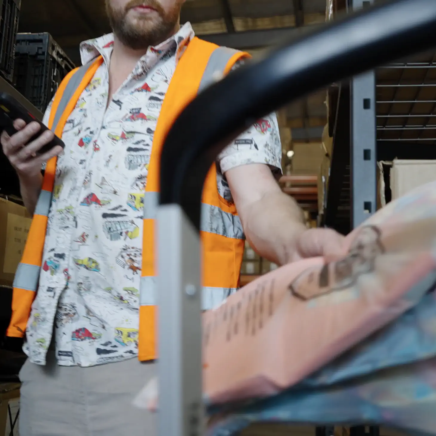 Warehouse worker wearing an orange safety vest holding a mobile device and pushing a cart with packages.