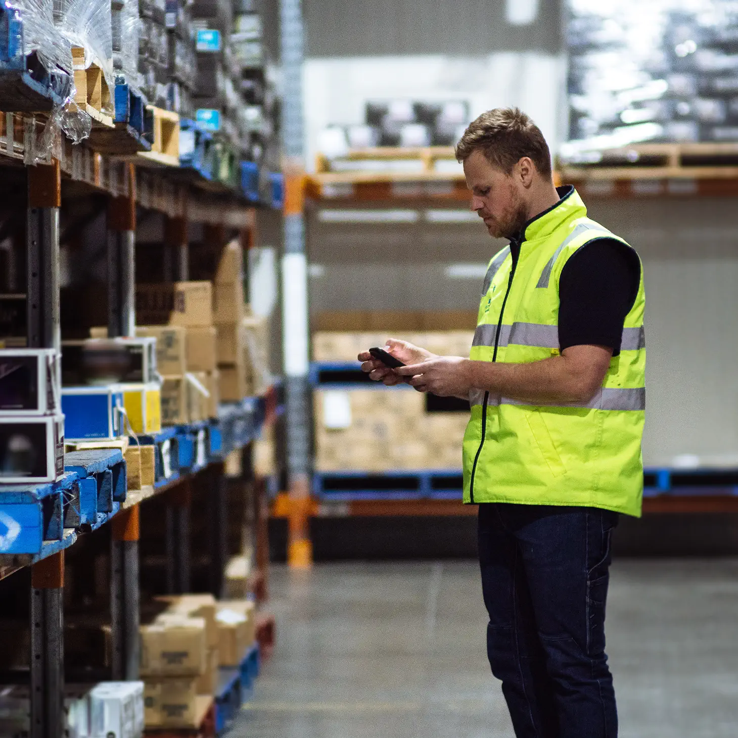 Warehouse worker in a neon yellow safety vest using a smartphone while standing next to shelves with boxed goods.