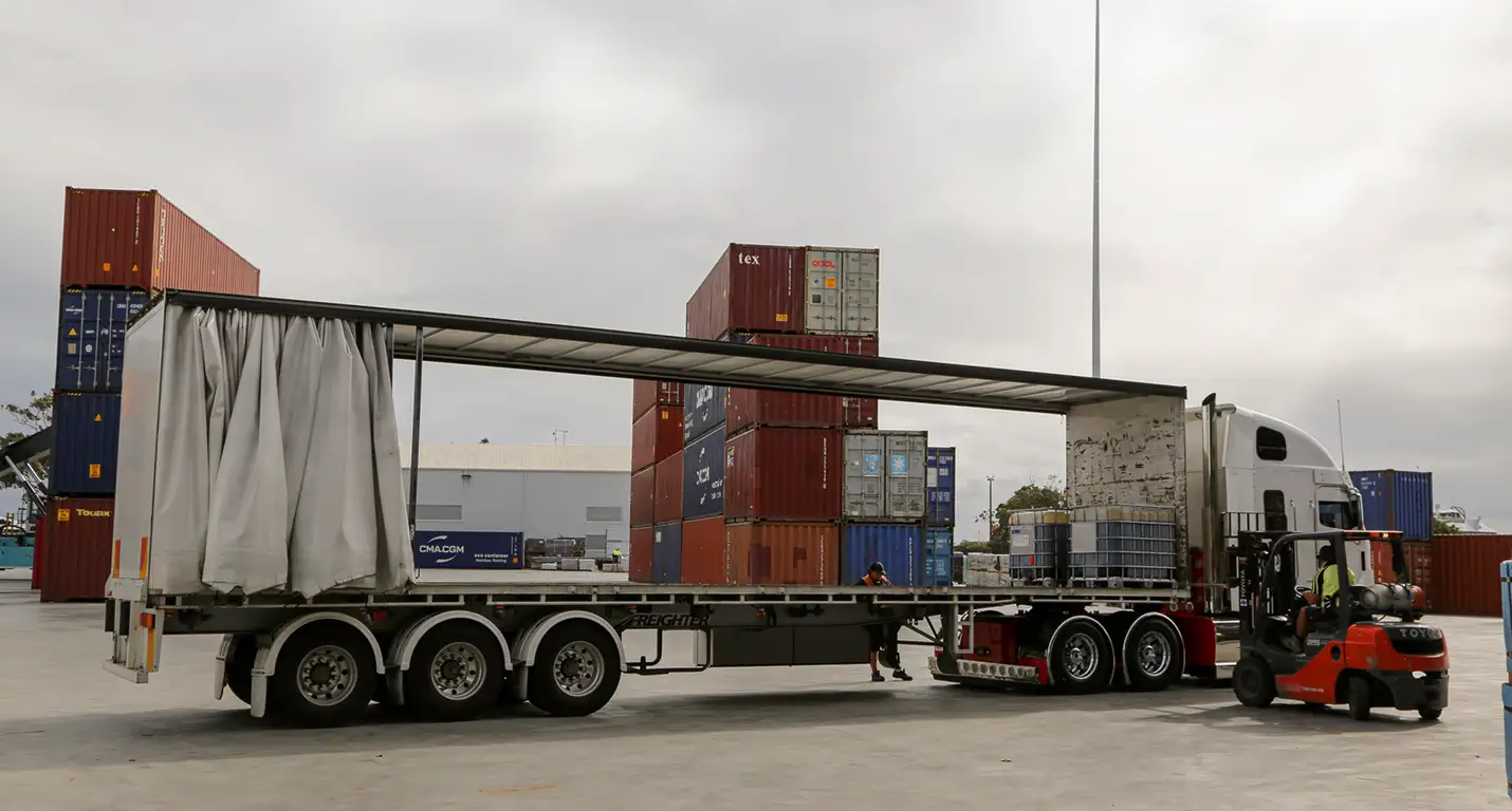 Truck with open side curtain being loaded with large containers by a forklift in a shipping yard stacked with cargo containers.
