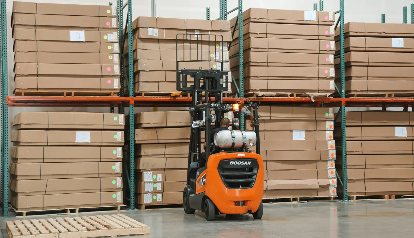 Forklift operated by a worker moving near tall stacks of large cardboard boxes on warehouse shelves.