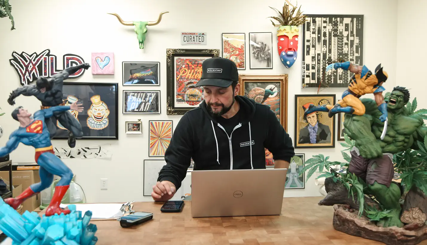 Man wearing Rockstar cap and hoodie working on a laptop at a desk, surrounded by superhero figurines and eclectic wall art.
