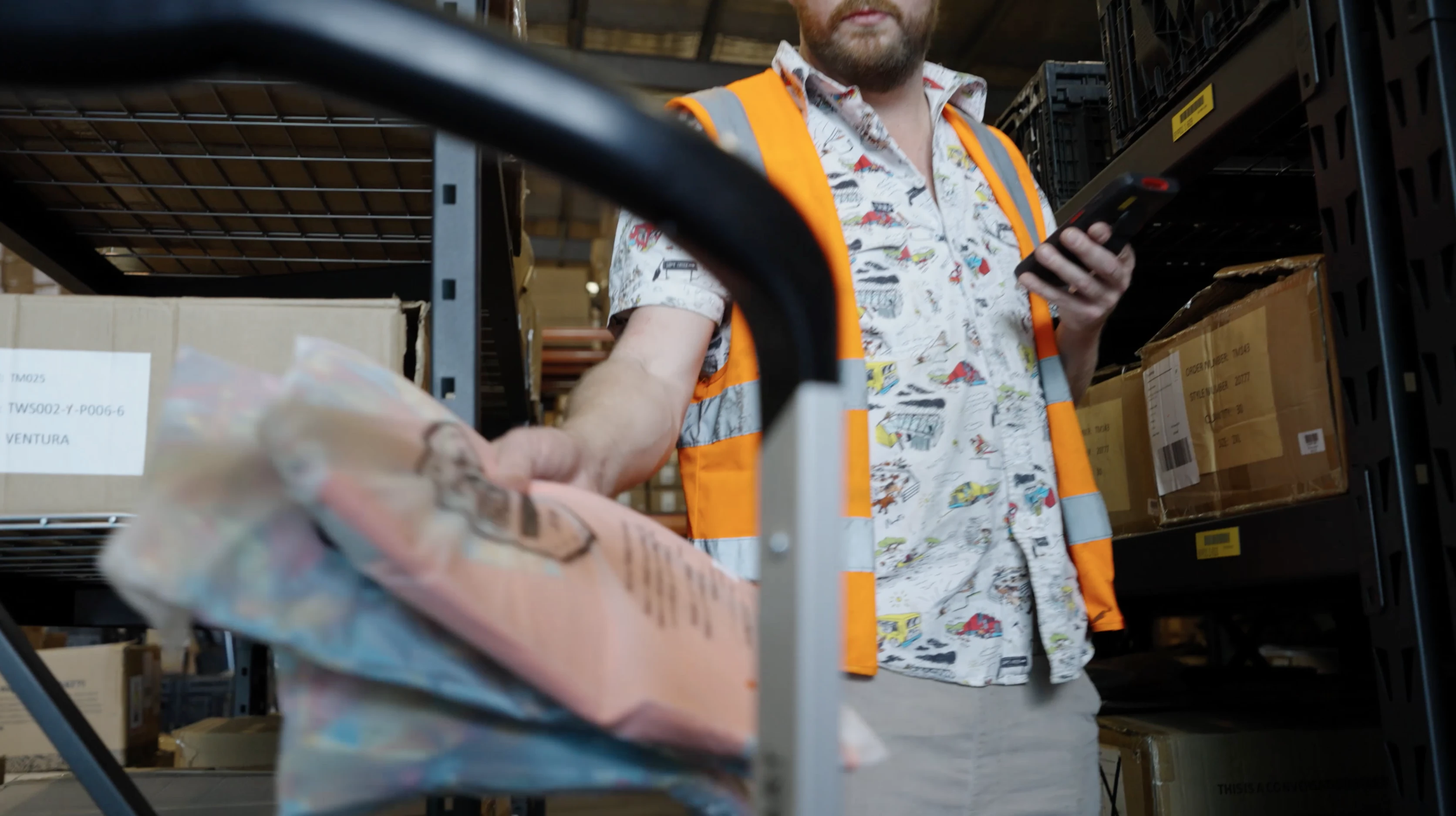 Warehouse worker in an orange safety vest using a handheld scanner and placing packaged items on a cart.