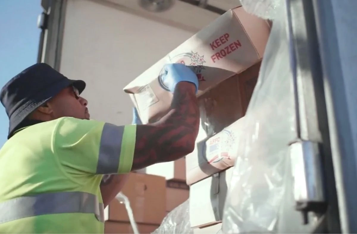 Worker in a high-visibility shirt and bucket hat unloading boxes labeled 'KEEP FROZEN' from a truck.