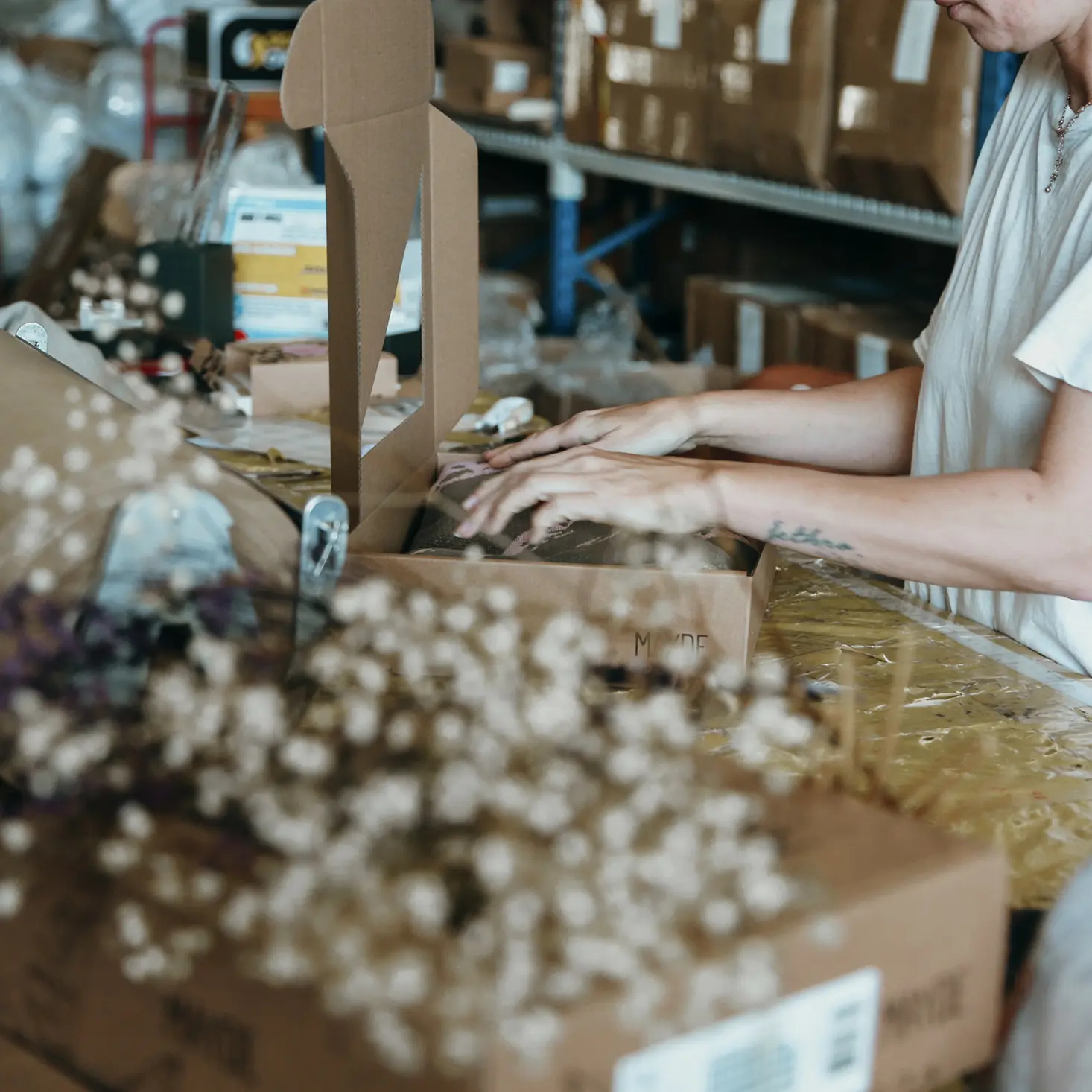 Person packing an item into a cardboard box on a cluttered worktable in a warehouse with shelves and boxes in the background.