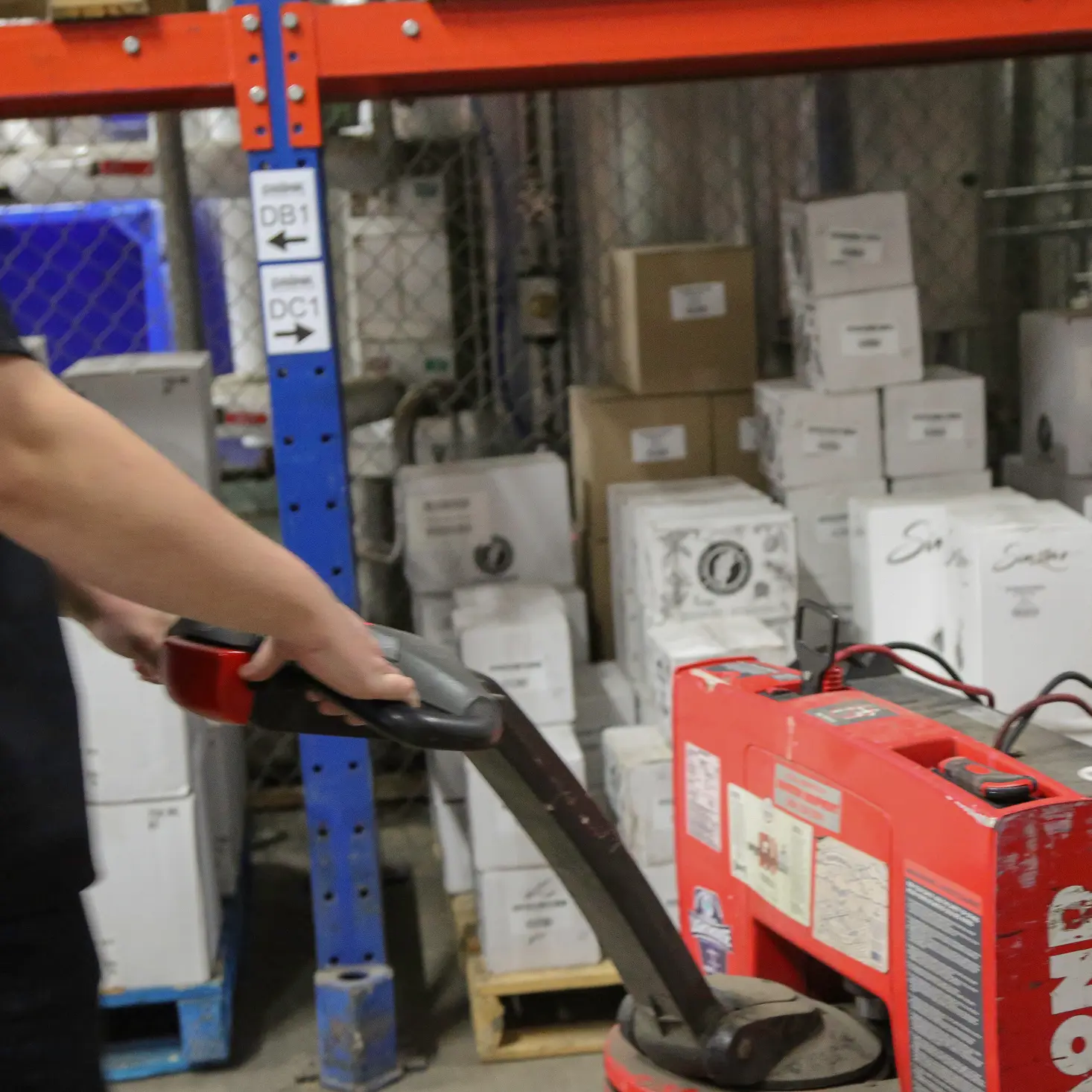 Person operating a red pallet jack in a warehouse with stacked boxes and metal racks.