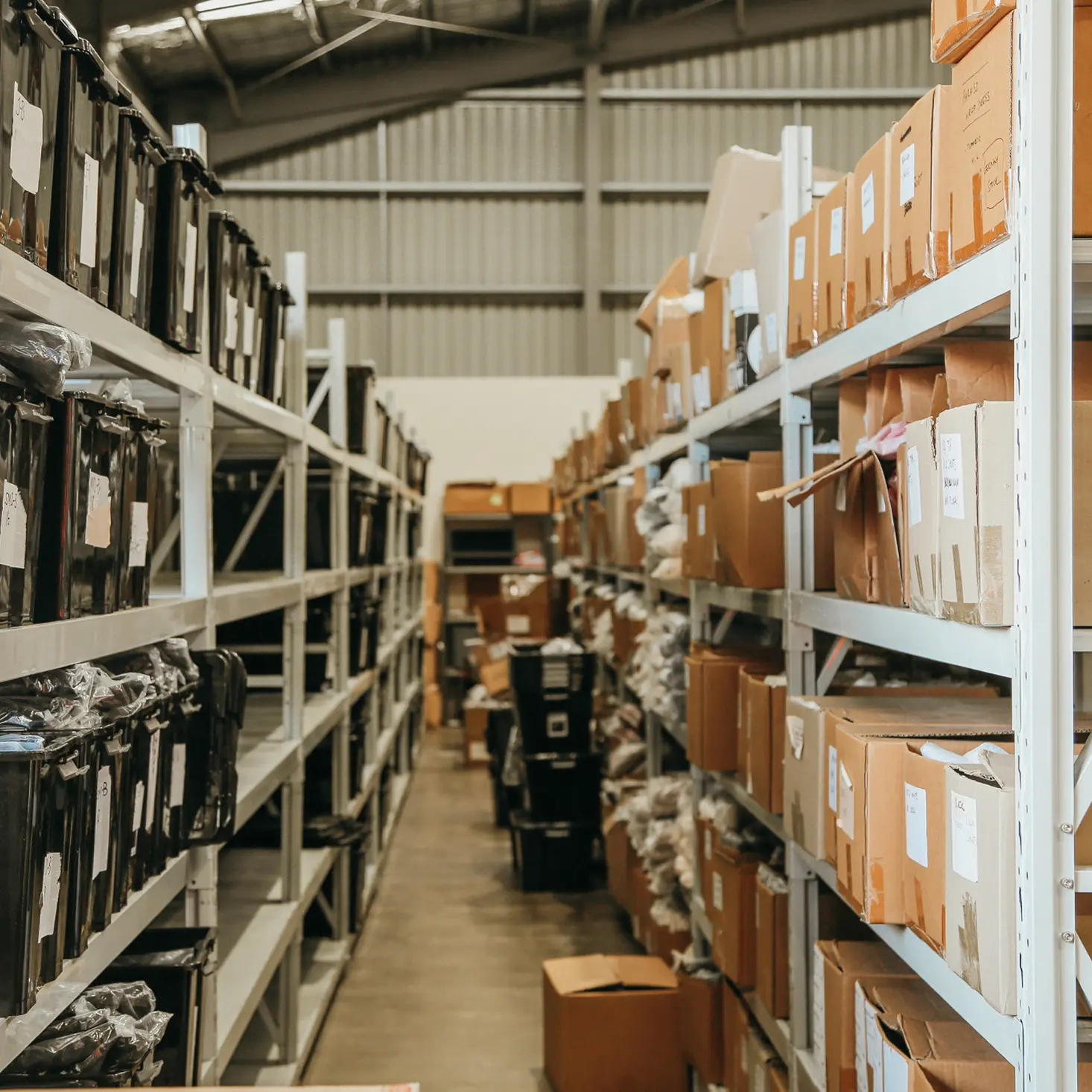 Warehouse aisle with metal shelves stocked with labeled black containers on the left and cardboard boxes on the right.