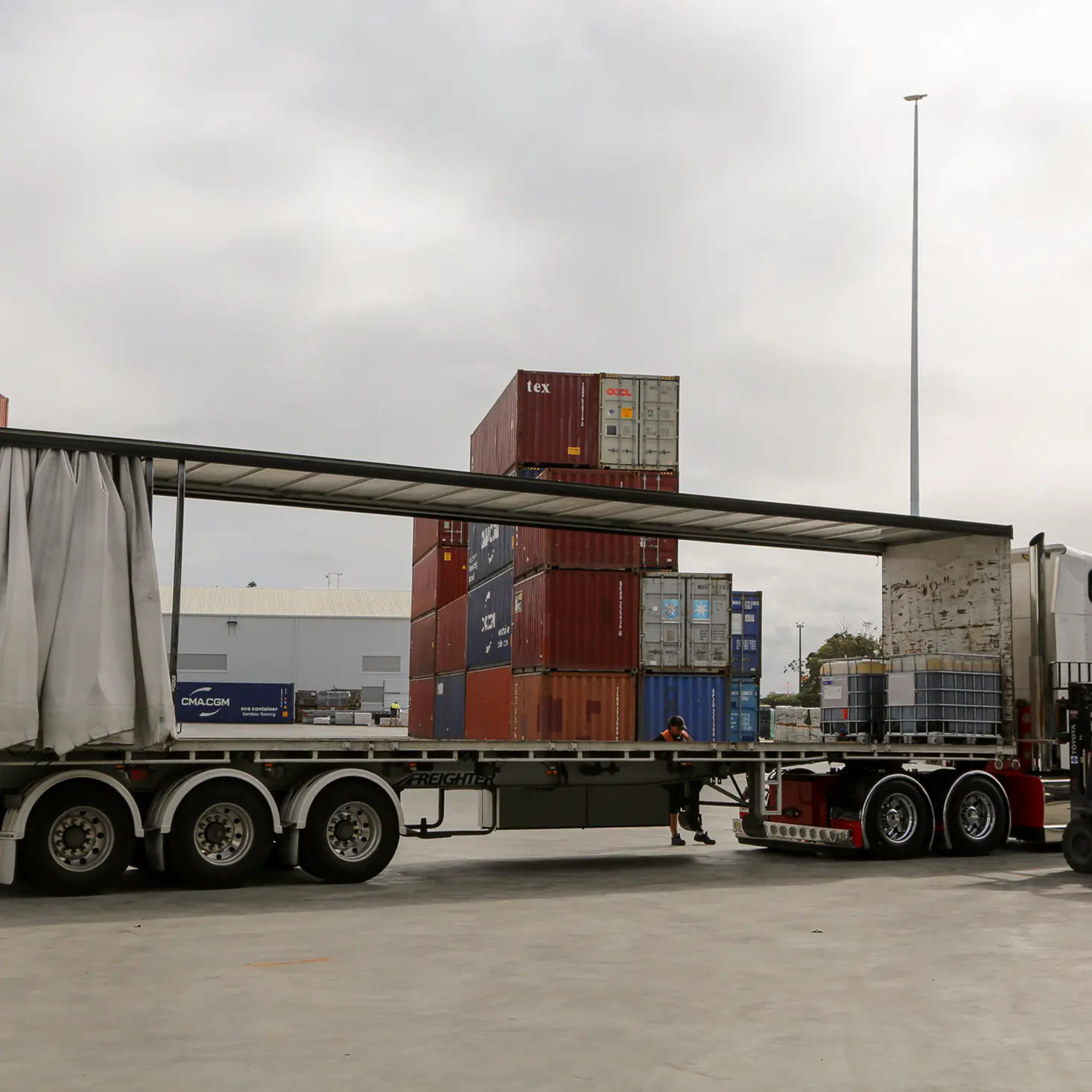 Flatbed semi-trailer truck with sides open parked in an industrial area with stacked shipping containers and a forklift nearby.