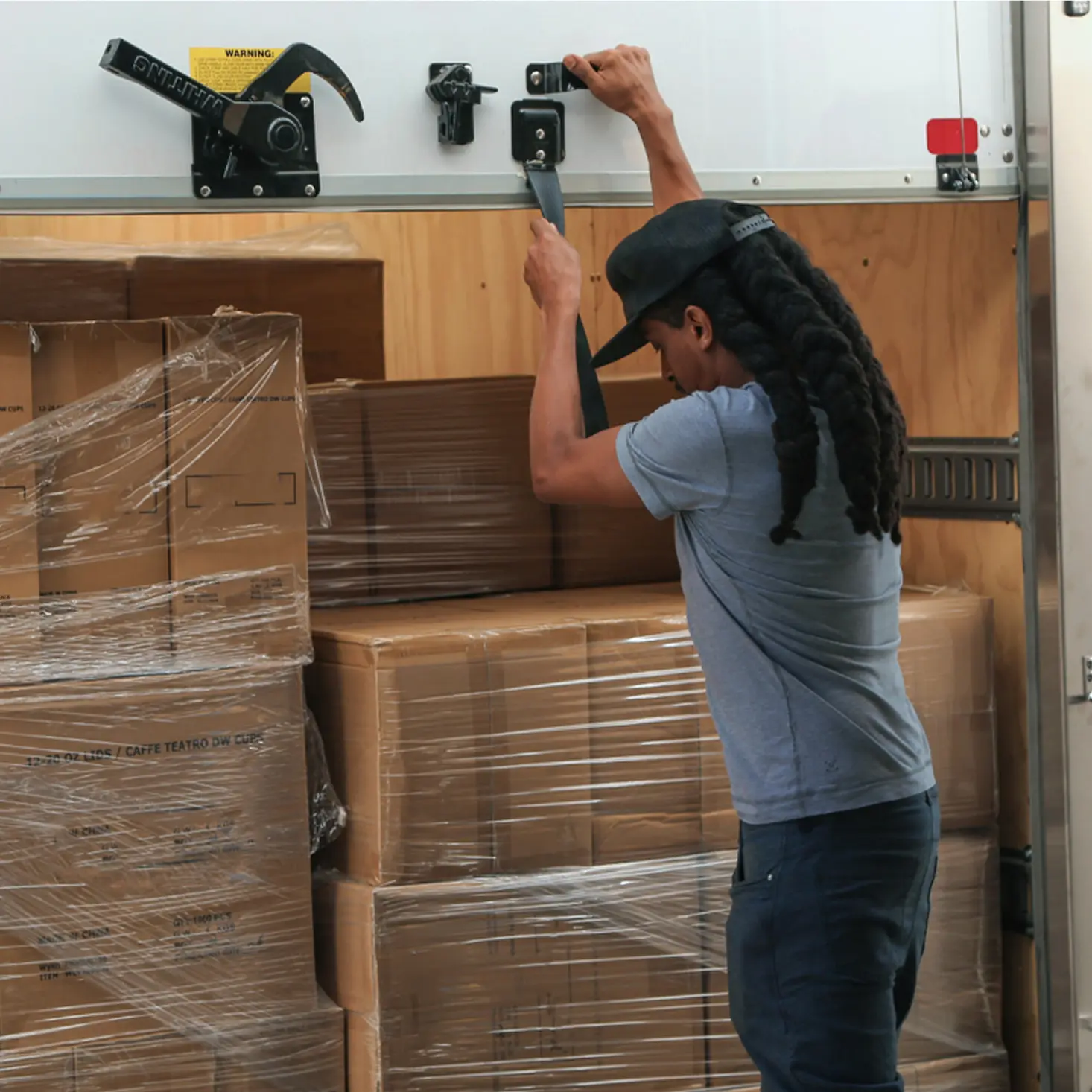 Man securing a strap inside a truck loaded with stacked cardboard boxes wrapped in plastic.
