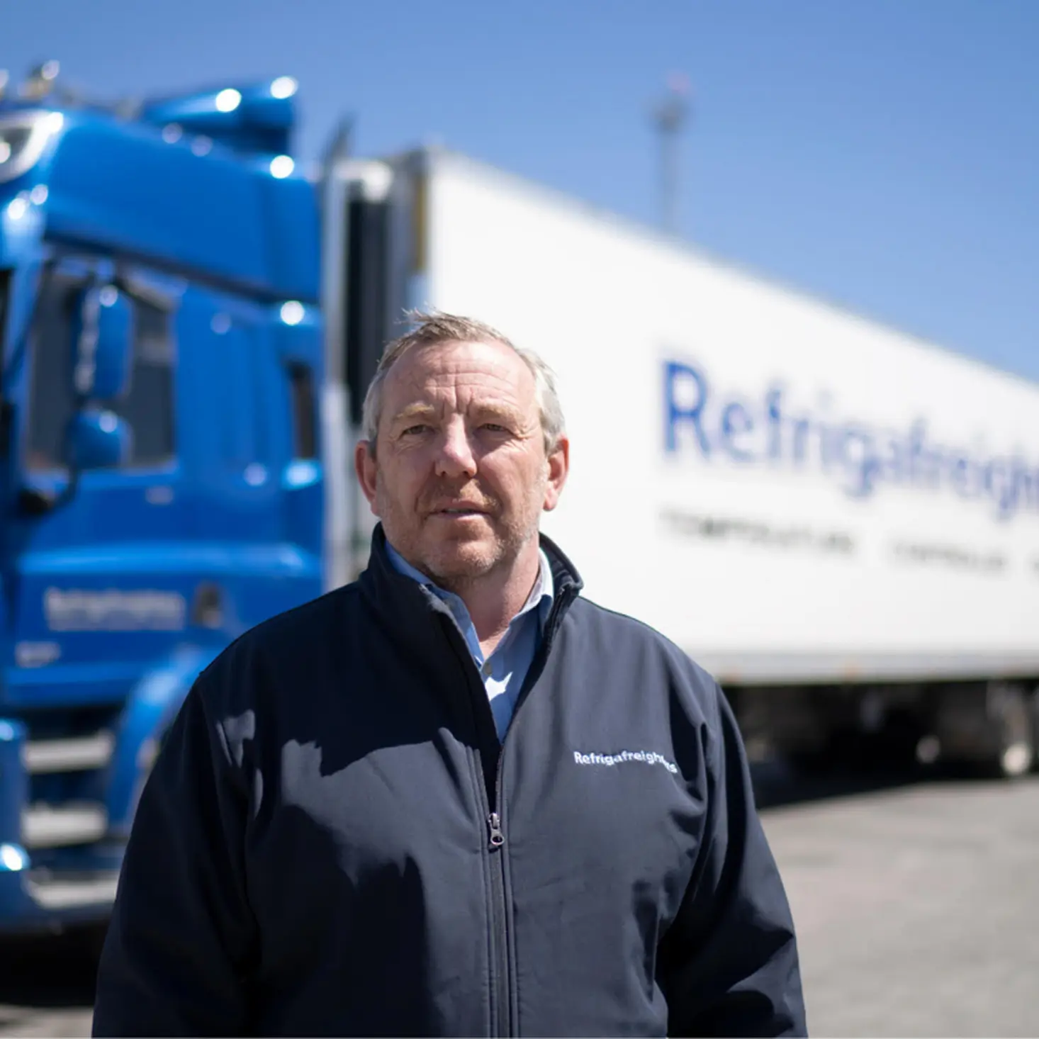 Man in a navy jacket standing in front of a blue truck and a white refrigerated trailer under a clear blue sky.